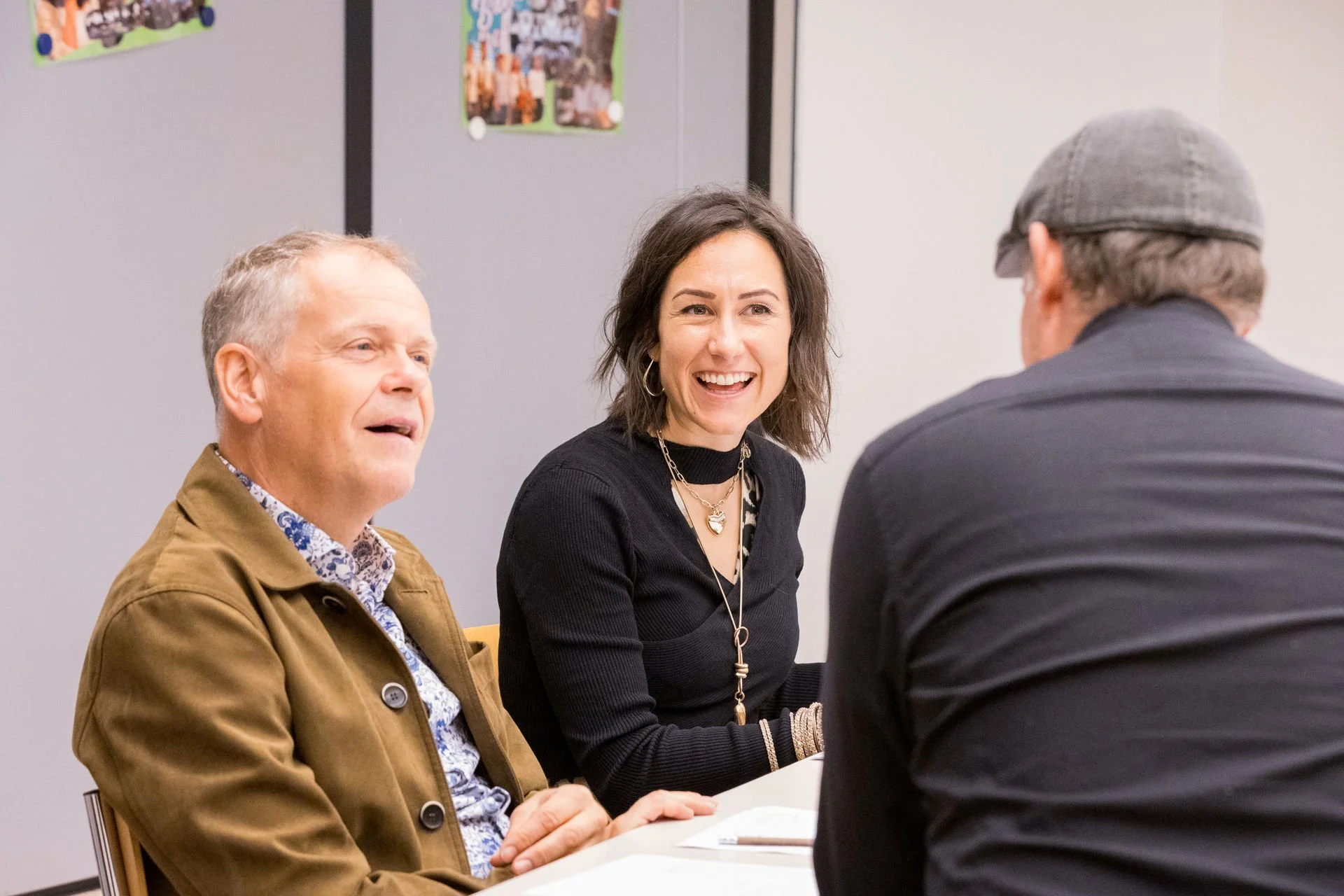 Drei Menschen sitzen an einem Tisch in einem Gespräch, die Frau in der Mitte lächelt freundlich, während die Männer auf beiden Seiten in die Richtung schauen.