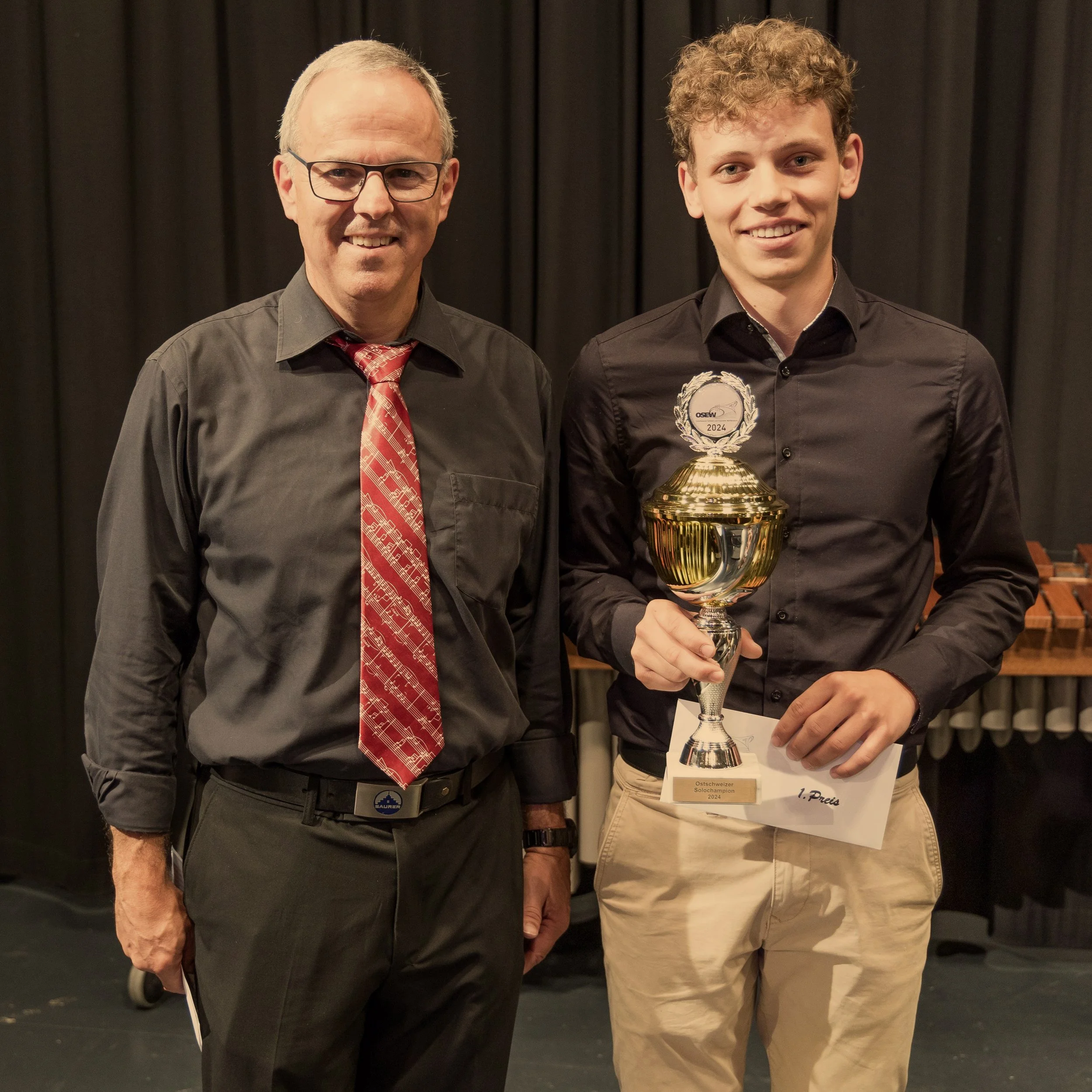 Zwei Männer stehen auf einer Bühne, der älteste trägt eine schwarze Hemd mit rotem Krawatte, der jüngere hält einen goldenen Pokal und eine Urkunde, beide lachen und posieren für das Foto.