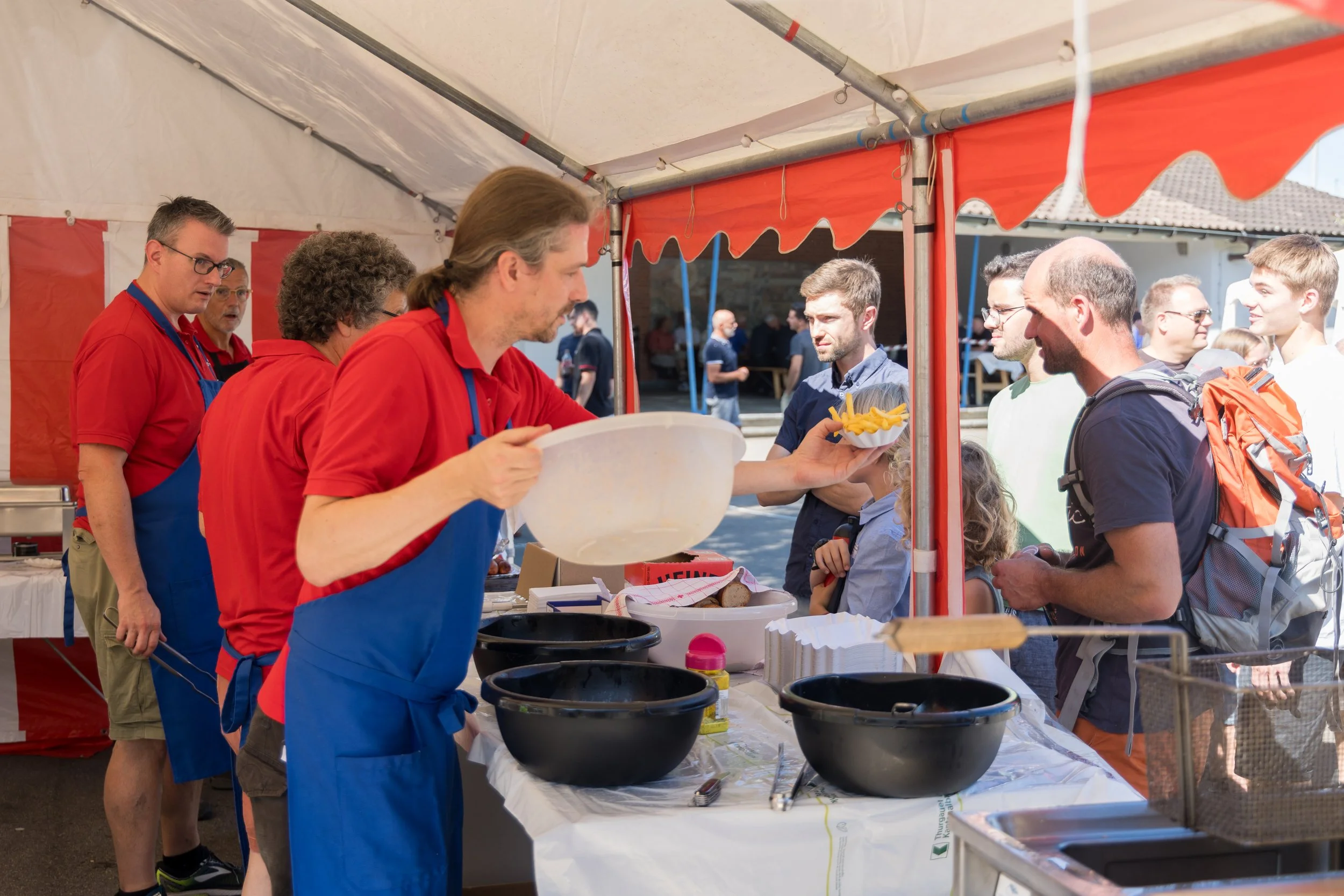 Menschen stehen an einem Essensstand, beim Essen bei einem Outdoor-Fest, mit einem Zelt und anderen Gästen im Hintergrund.