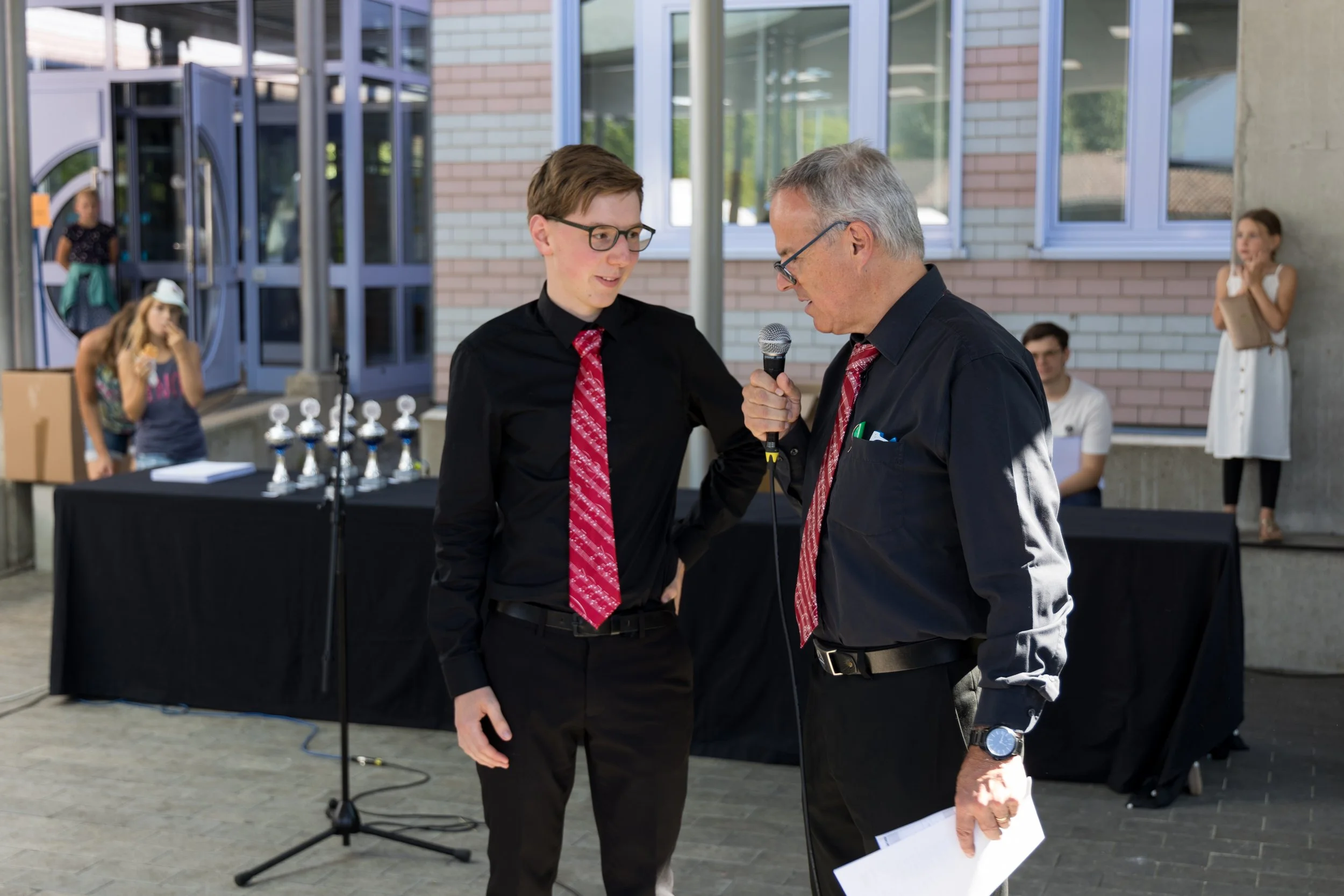 Ein älterer Mann im schwarzen Hemd und roter Krawatte spricht mit einem jungen Mann in schwarzem Hemd und roter Krawatte. Im Hintergrund stehen Trophäen auf einem Tisch. Es scheint eine Preisverleihung oder Zeremonie zu sein.