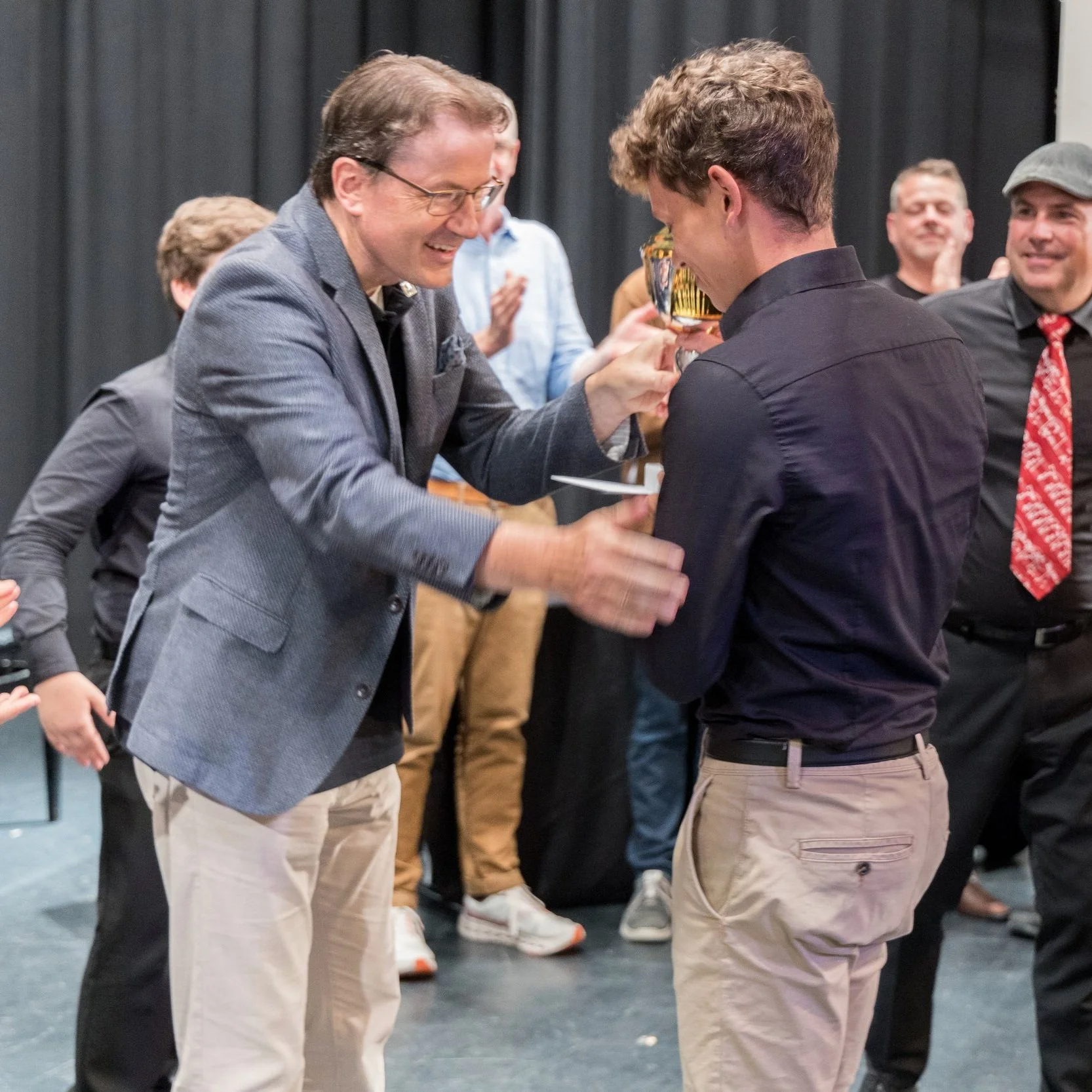Ein Mann in grauem Anzug überreicht einem jüngeren Mann in schwarzem Hemd eine Auszeichnung, während andere Menschen im Hintergrund applaudieren.