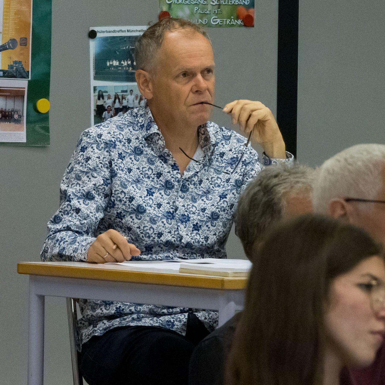 Mann mit Brille in Hemd mit Blumenmuster, sitzt an einem Tisch, hält die Brille in der Hand und sieht nach vorn, im Hintergrund ein schwarzer Kasten und Poster an der Wand.