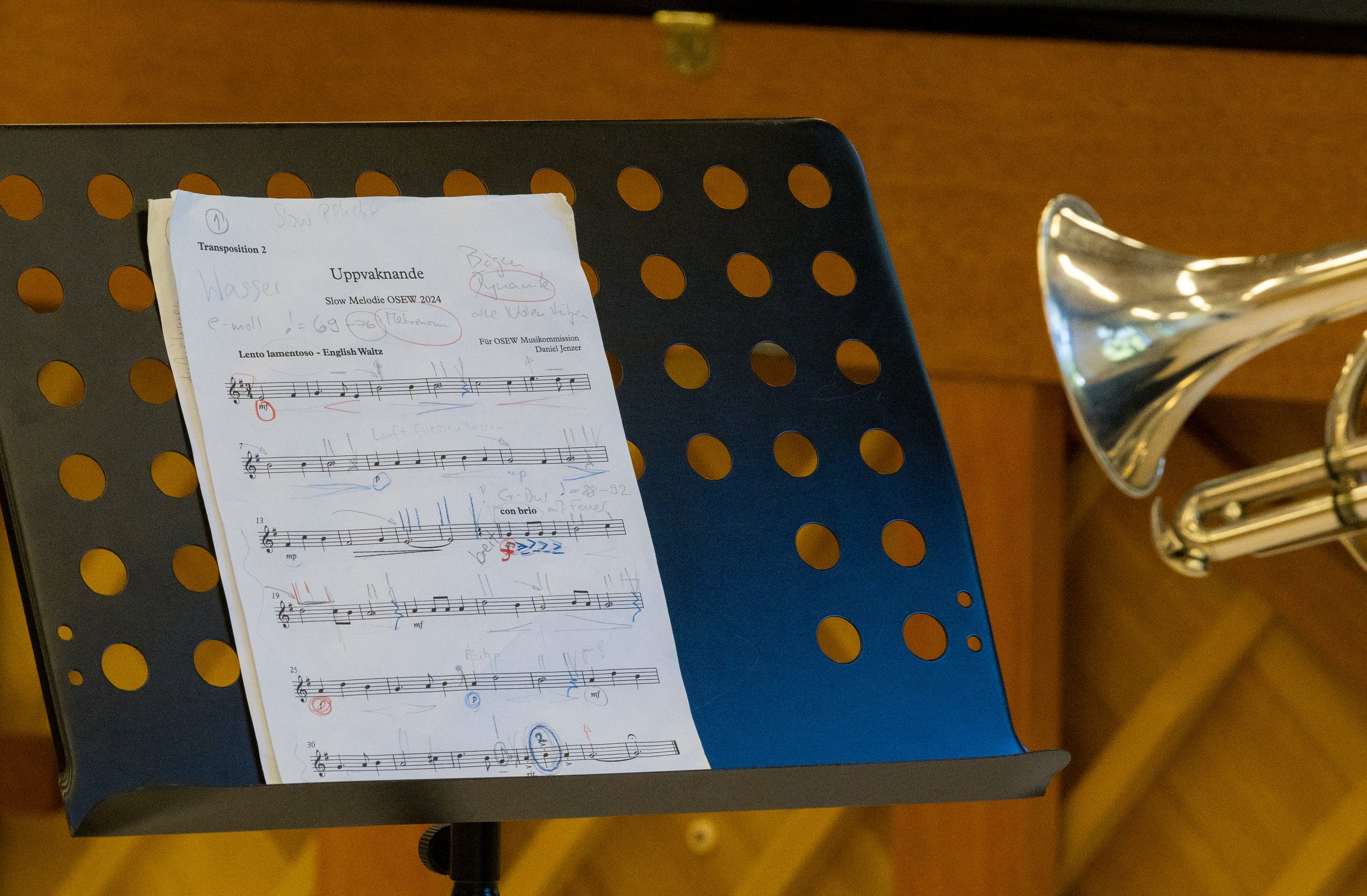 Notenblatt auf einem Musikständer mit einem Blasinstrument links neben dem Ständer, vermutlich eine Trompete, im Hintergrund ein Holzfußboden.