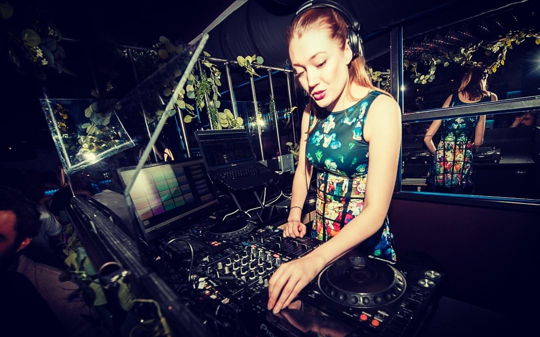 Kvinne som er DJ og spiller musikk ved en miksebord i en klubbsetting, omringet av planter og speil.