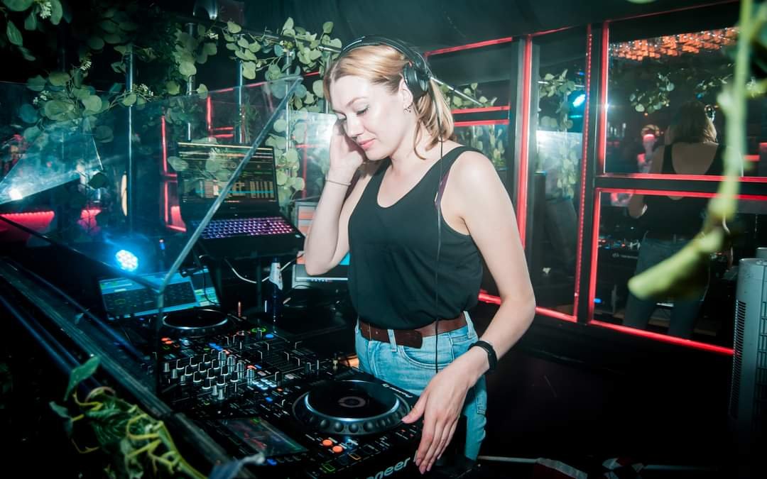 Kvinne i svart topp bruker DJ-utstyr i en nattklubb med grønn og rød belysning.