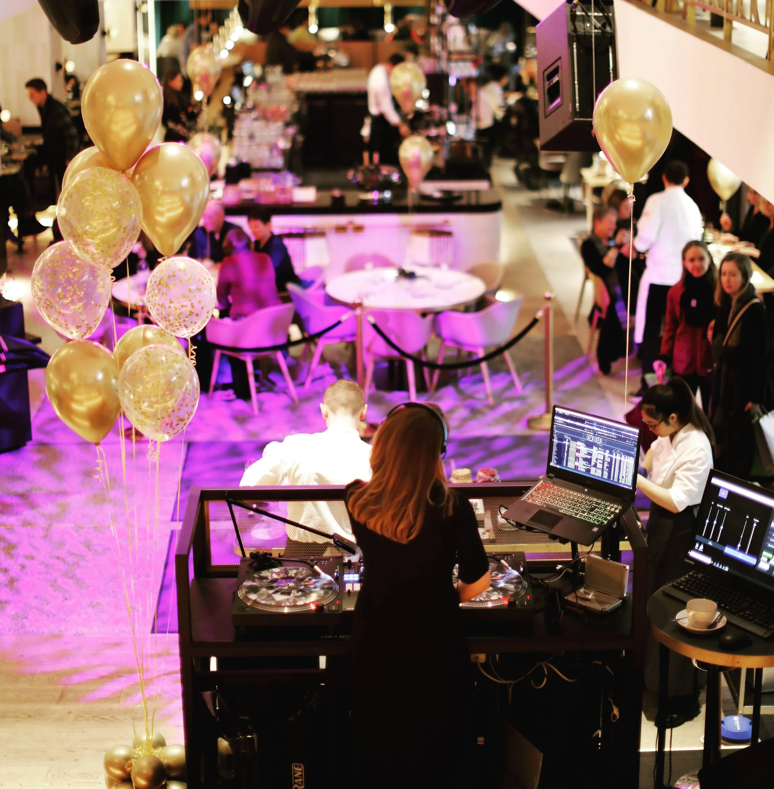 En DJ spiller musikk på en fest eller arrangement i en restaurant eller kafé. Det er ballonger og mennesker som sitter ved bordene og sosialisere. Atmosfæren er livlig og belyst med lilla lys.