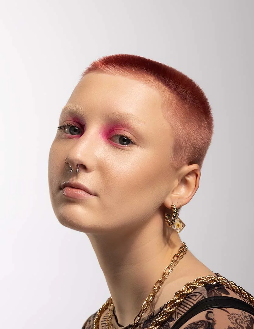 A person with short, reddish hair, wearing multiple gold necklaces and earrings, has pink eyeshadow, light skin, and a septum piercing, posing against a plain light background.