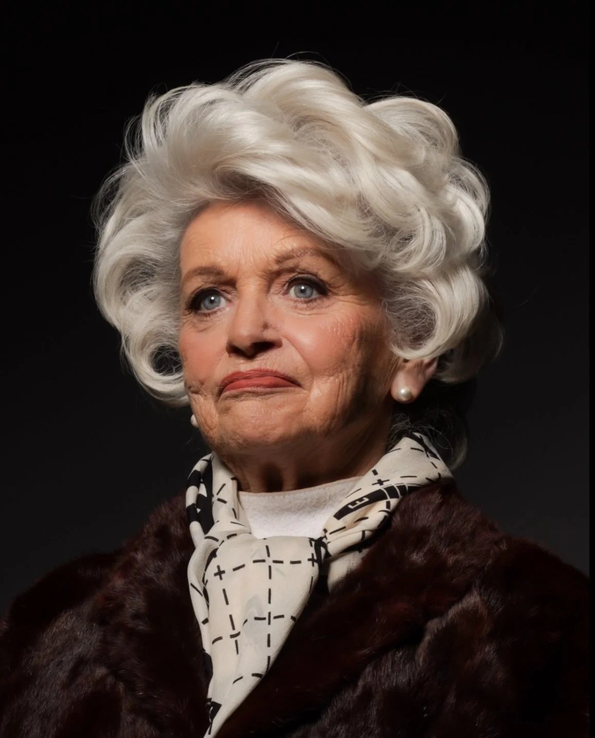 An elderly woman with styled white hair, blue eyes, wearing pearl earrings, a patterned scarf, and a dark fur coat against a black background.