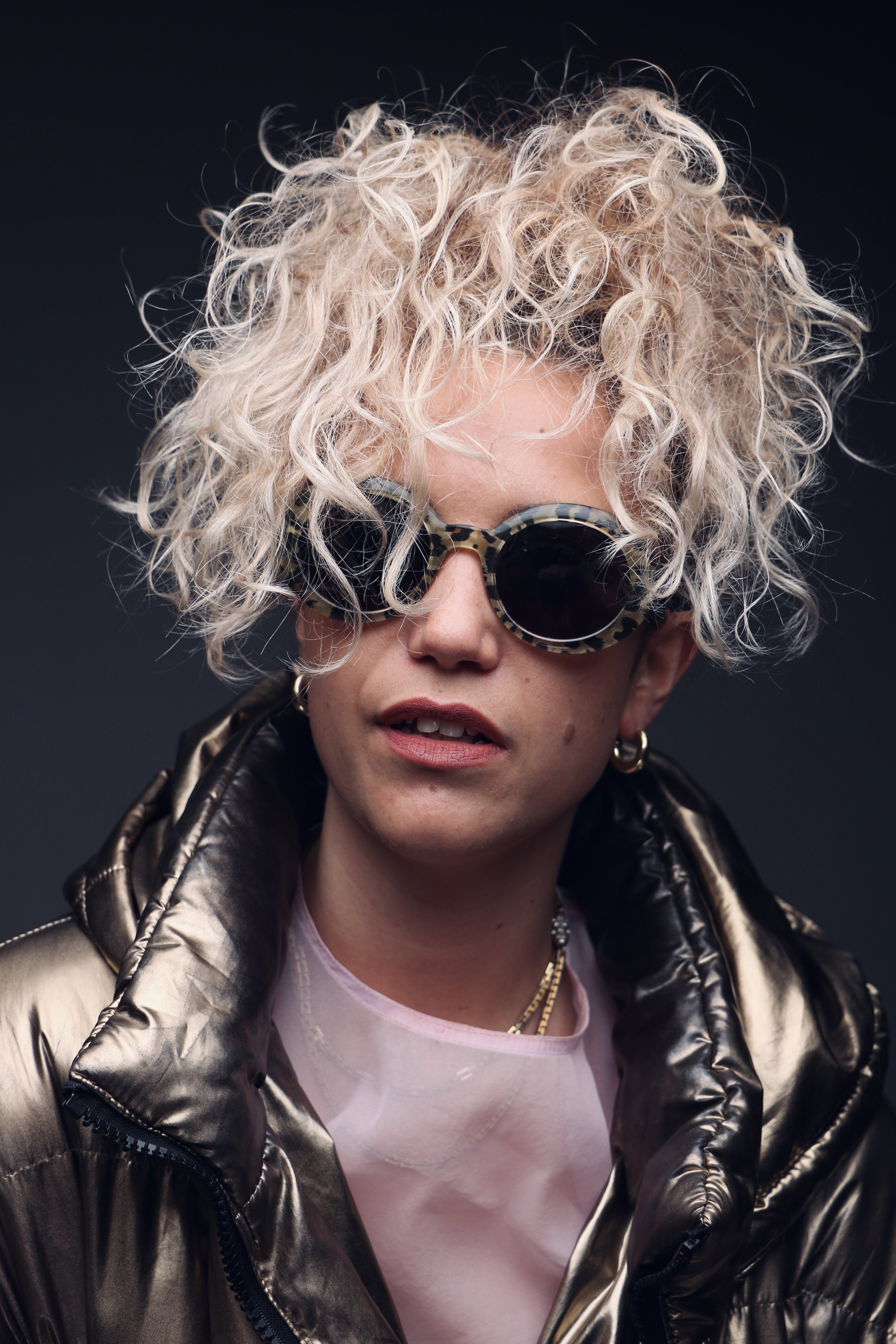 A person with curly platinum blonde hair wearing oversized leopard-print sunglasses, a shiny black puffer jacket, a pink t-shirt, earrings, and layered necklaces against a dark background.