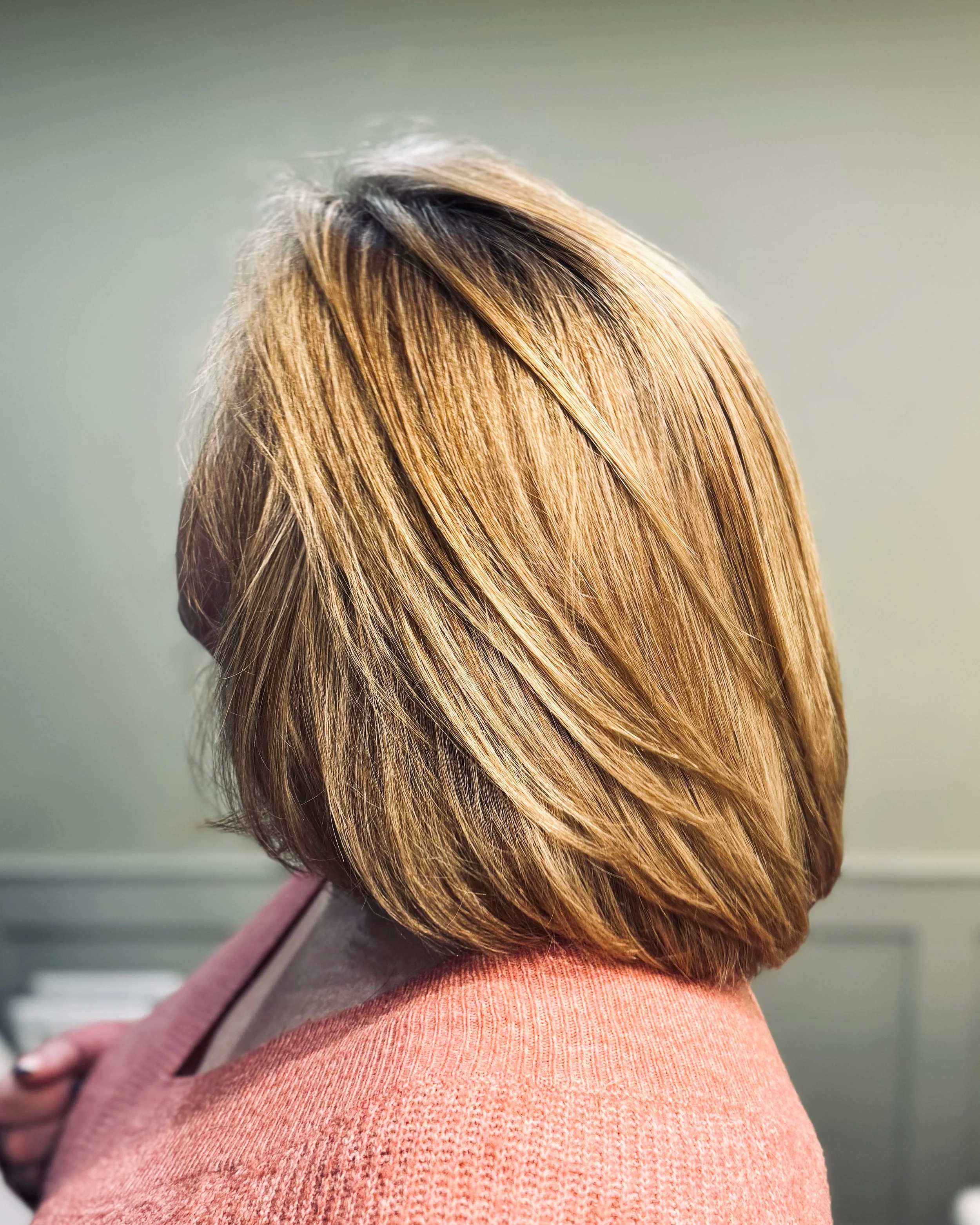 Back view of a woman with shoulder-length, layered blonde hair, wearing a pink sweater.