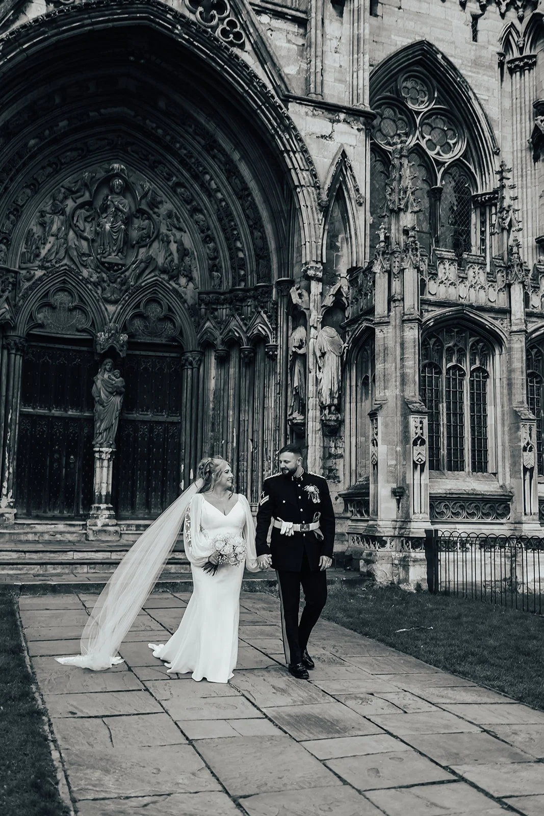 Wedding couple walking in front of a historic stone building with ornate arch and statues.