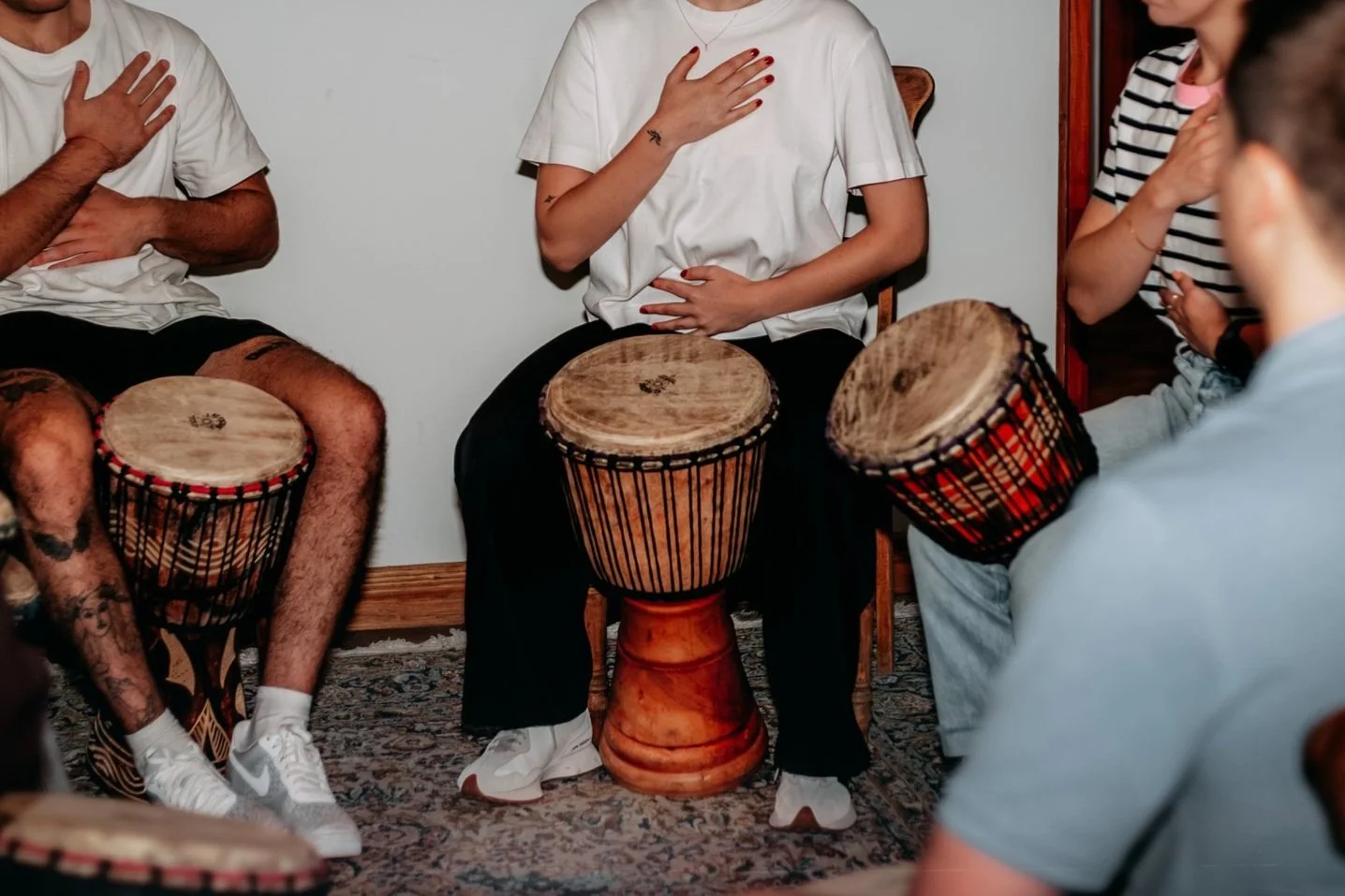 People sitting around in a circle, with djembe drums in front of them, hands on their chests.