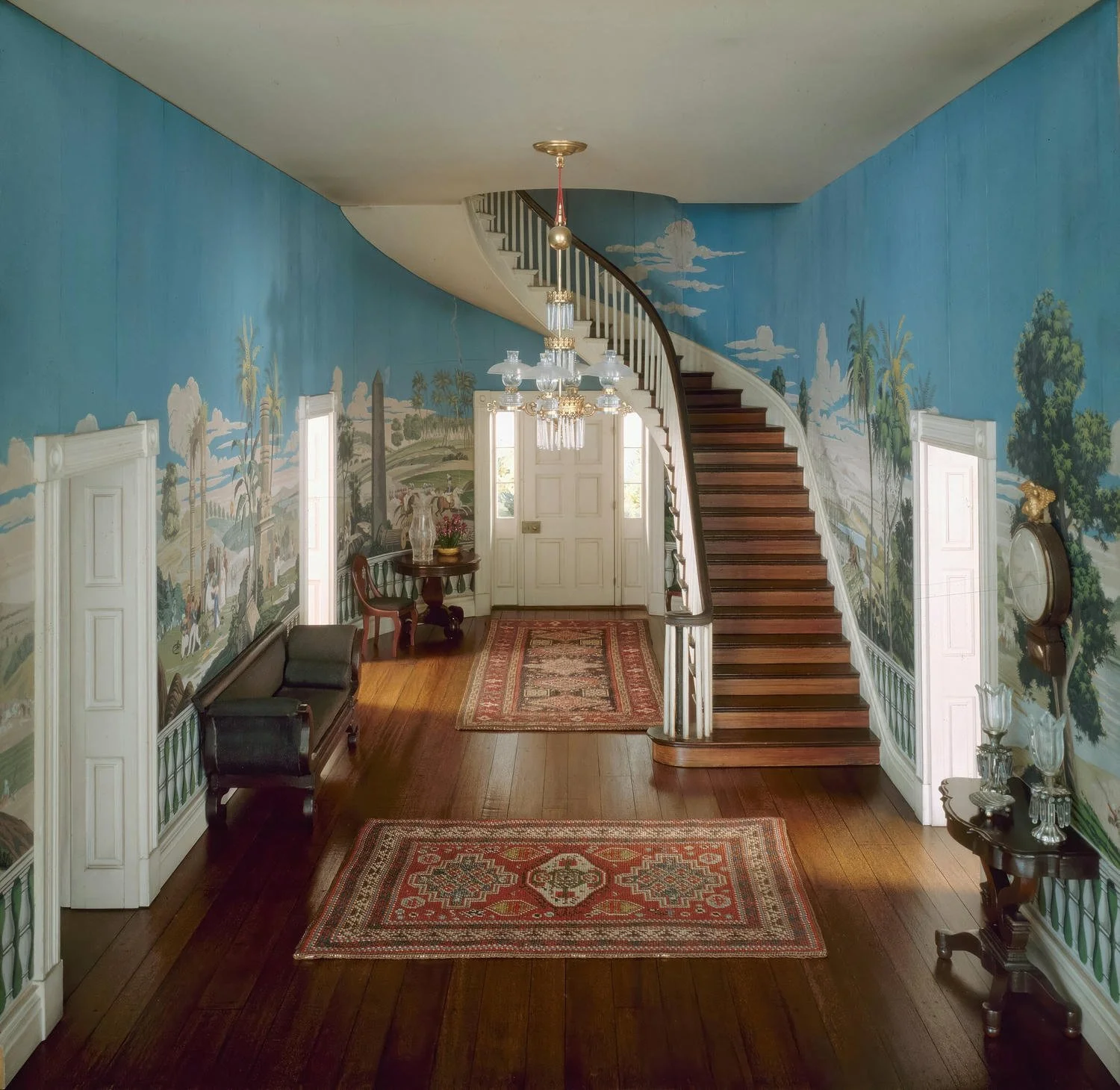 Interior of a historic mansion entry hallway, featuring a wooden staircase, framed landscape murals on blue walls, ornate ceiling chandelier, vintage furniture, and an area rug.