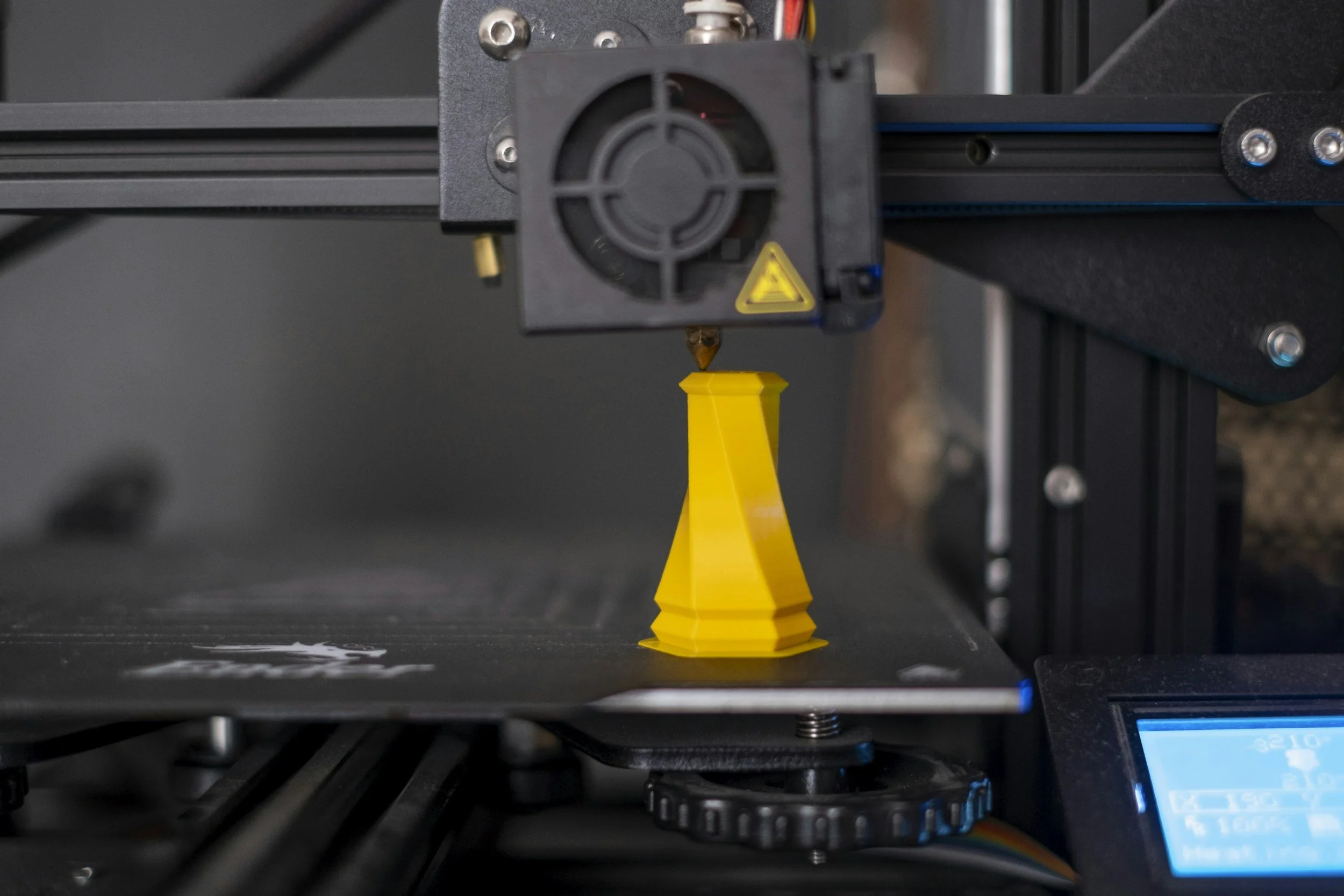 A 3D printer creating a yellow 3D printed object on the print bed with the printer's extruder nozzle in use.
