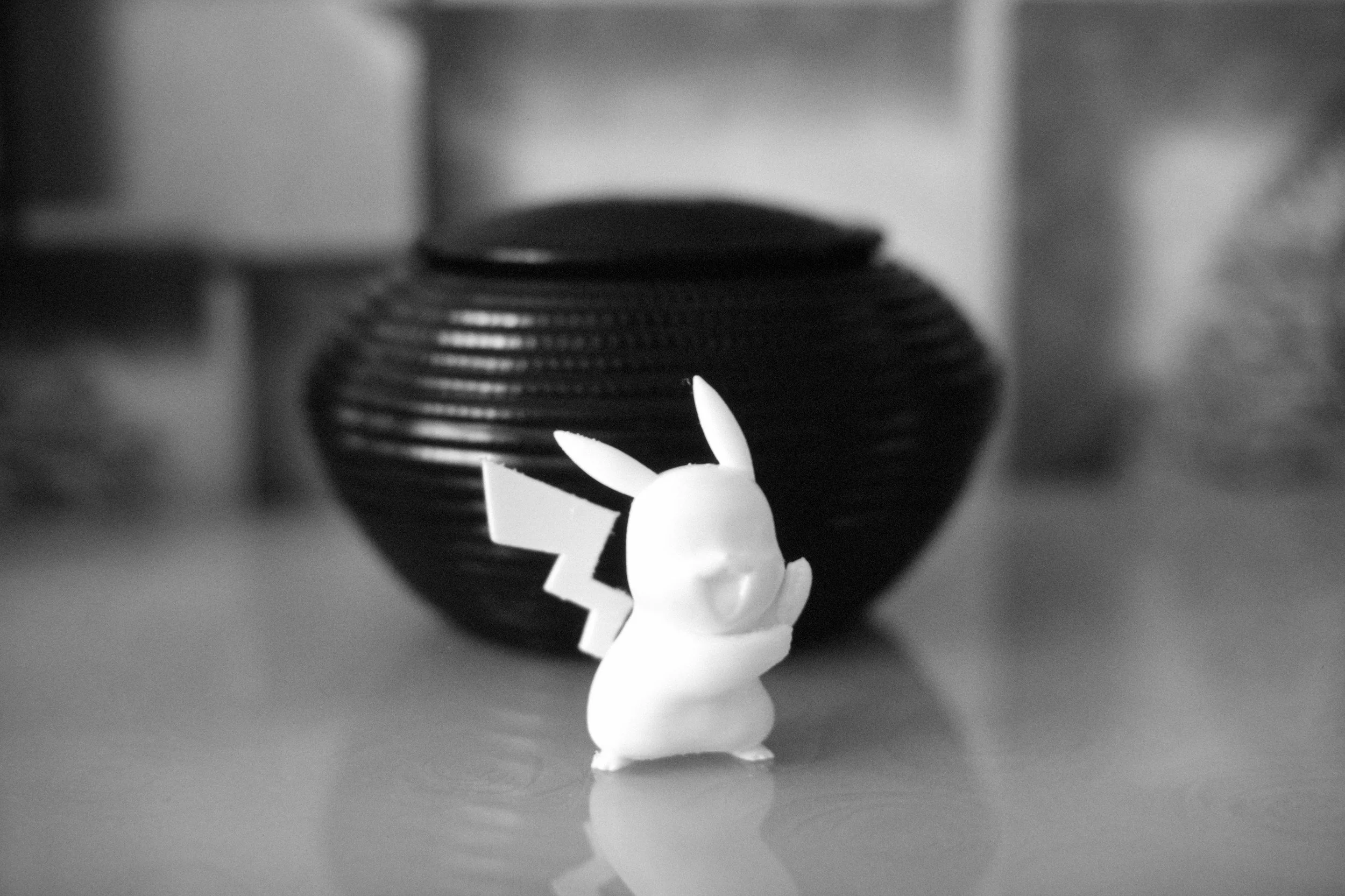 A small 3d printed Pikachu figurine facing away from a large, round, black textured vase on a wooden surface in black and white.