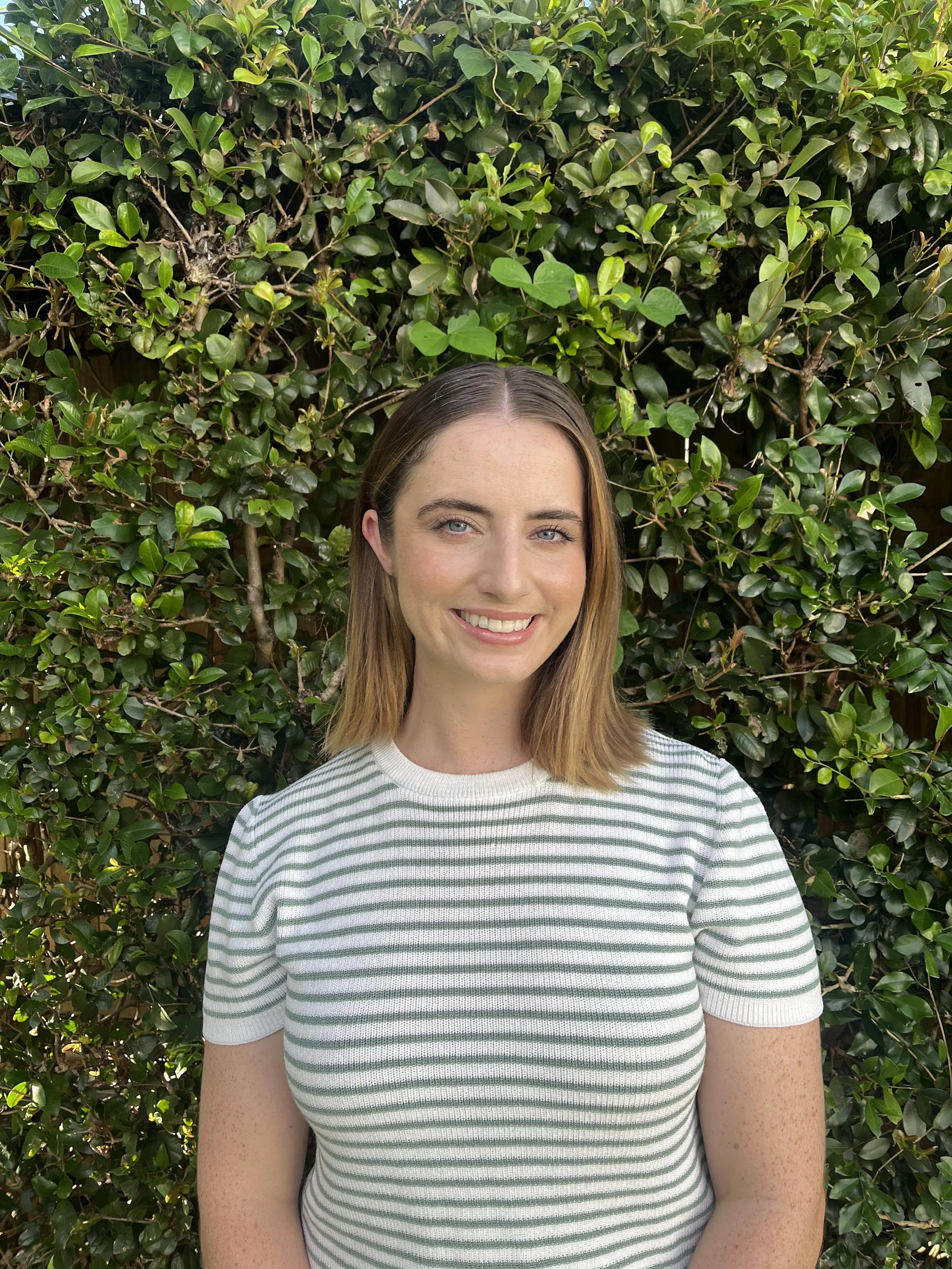 Smiling person with green striped shirt standing in front of a lush green hedge.