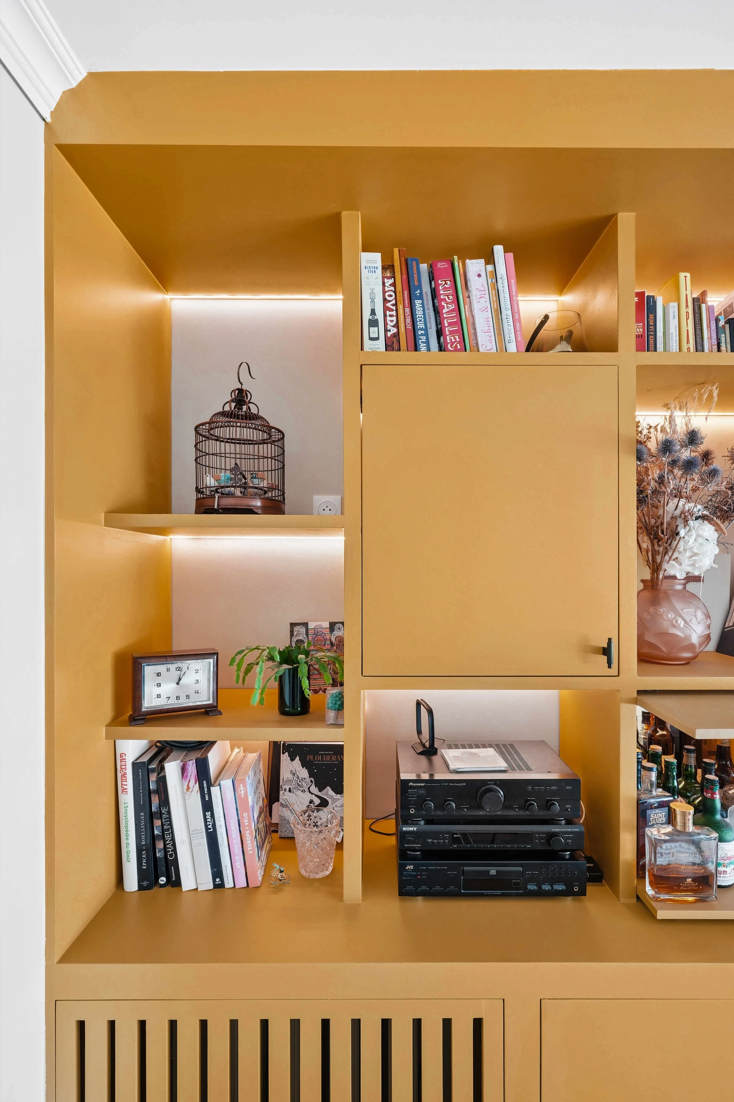 Détail d'une bibliothèque sur mesure ocre avec niches éclairées, rangements fermés et étagères ouvertes.