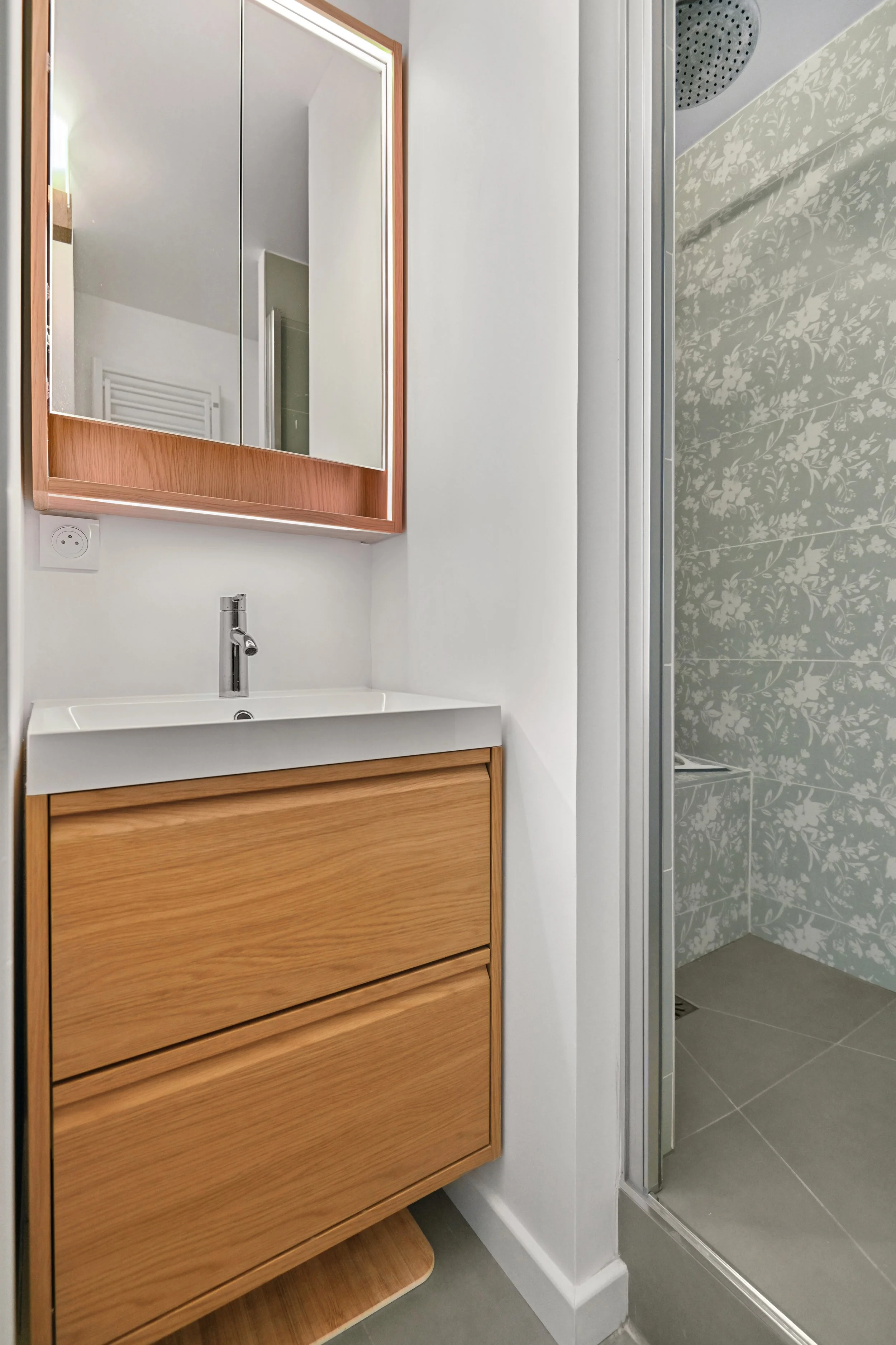 Salle d’eau rénovée avec meuble vasque en bois et douche aux motifs végétaux gris-vert.