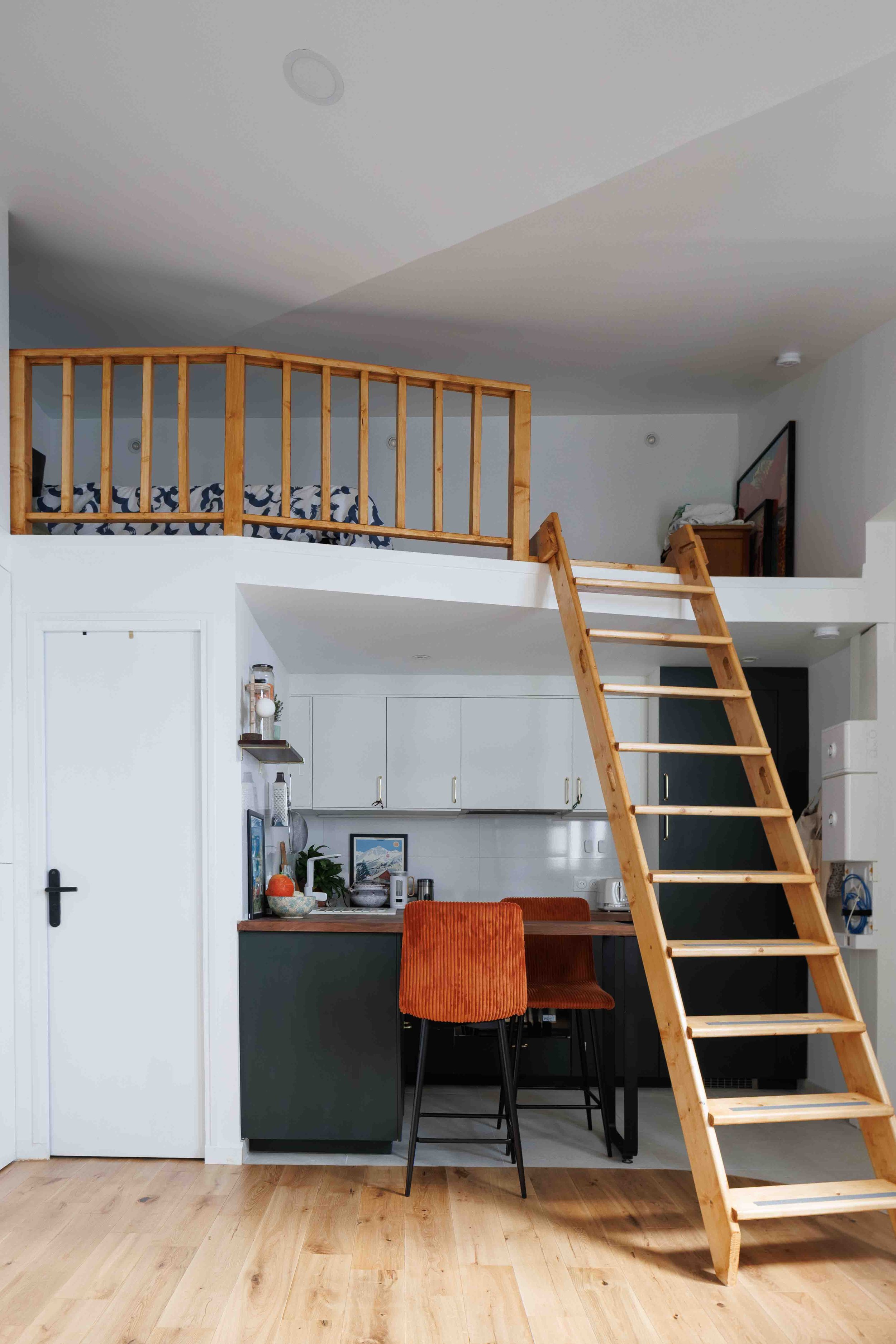 Vue plongeante sur l’espace mezzanine baigné de lumière naturelle, révélant une charpente rénovée et une décoration minimaliste aux teintes claires. Une échelle graphique donne accès à l’étage.