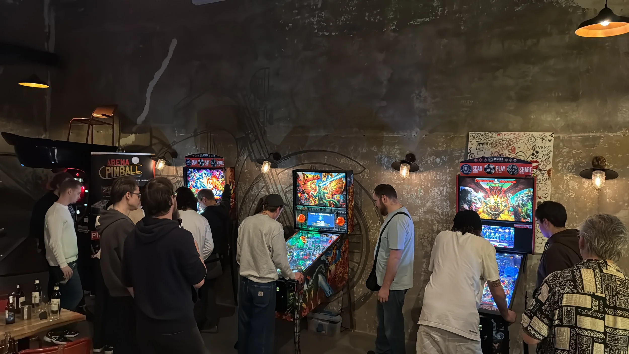 People playing pinball and arcade games in a dimly lit arcade with a dark, textured wall in the background.