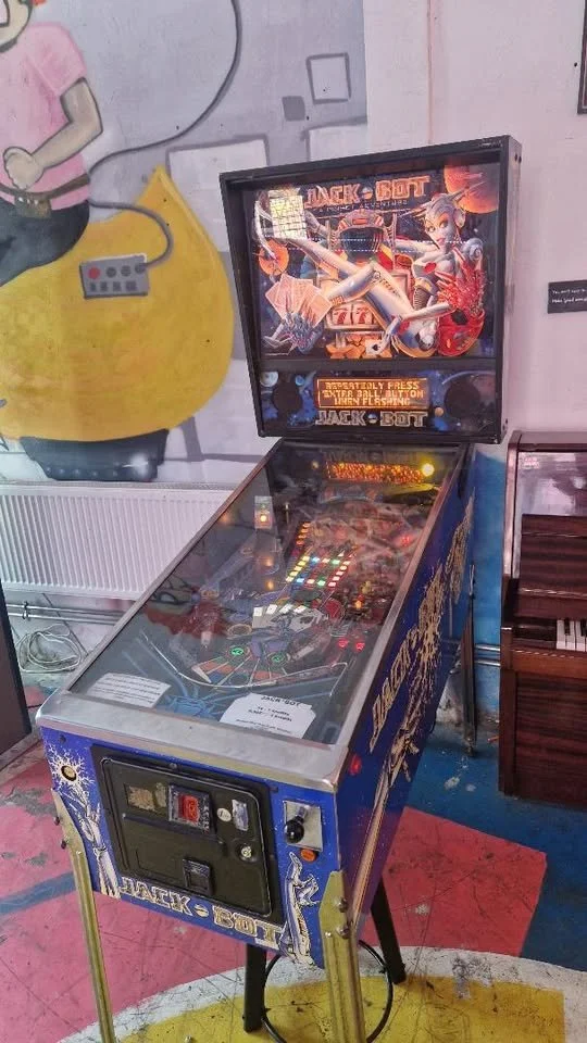 A vintage Jack-Bot pinball machine with colorful artwork on the backglass, featuring a robot girl, and game controls on the front, situated in an arcade with a mural on the wall behind.