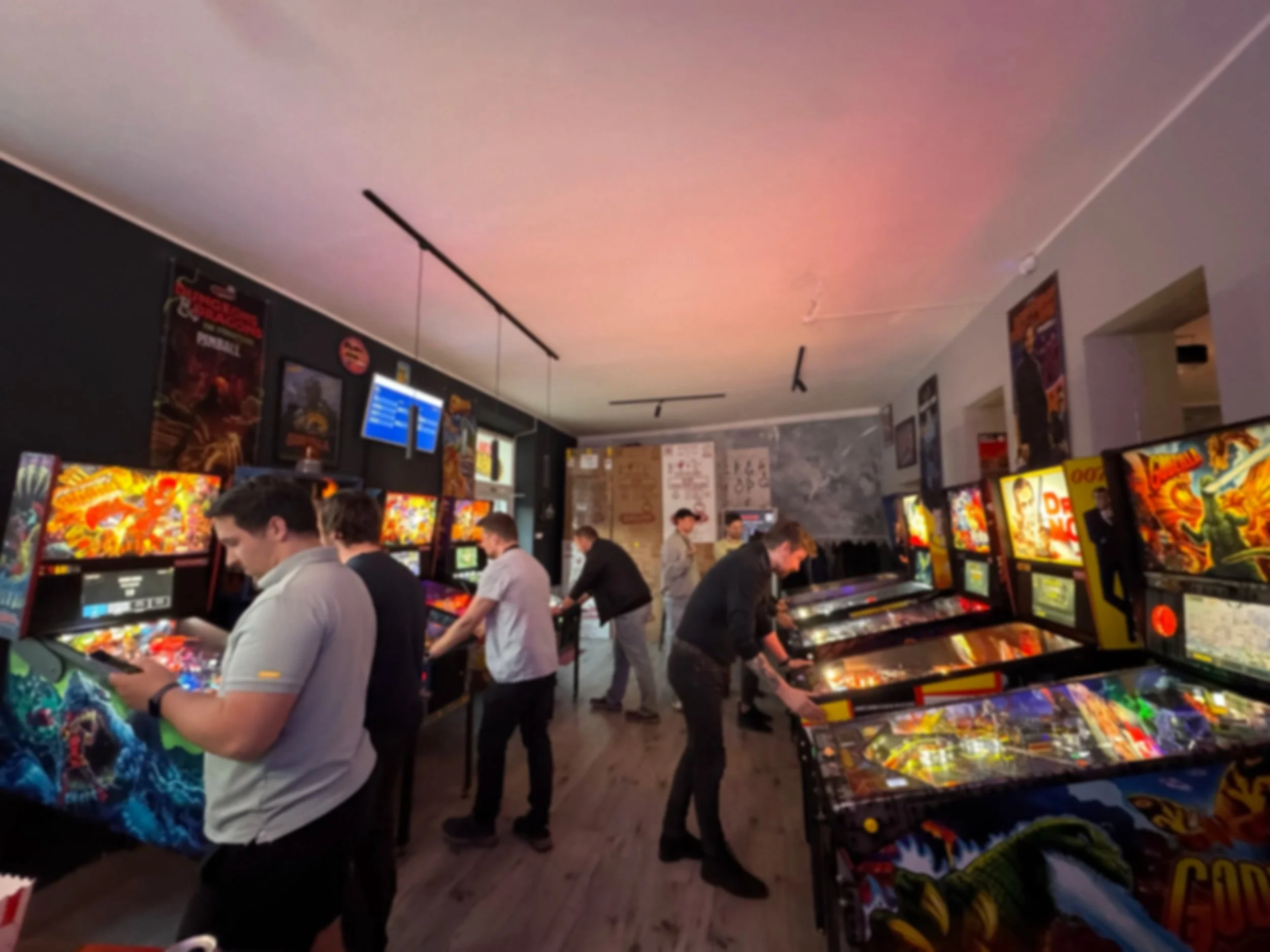 People playing pinball at an arcade with multiple pinball machines, posters on the wall, and a ceiling with track lighting.