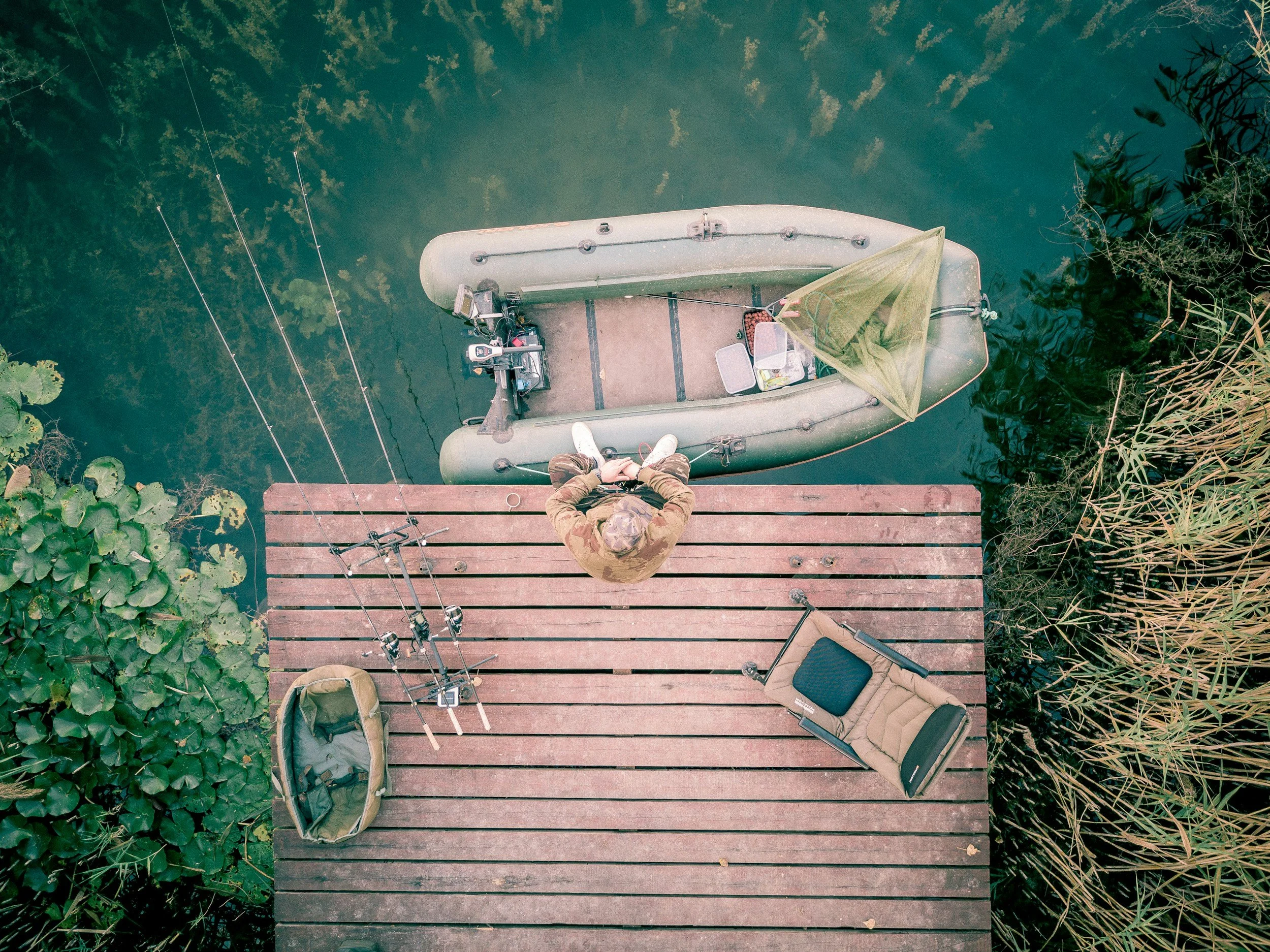 Overhead view of a person standing on a wooden dock next to a small boat with fishing rods, a tackle box, and a folding chair, surrounded by water and green vegetation.