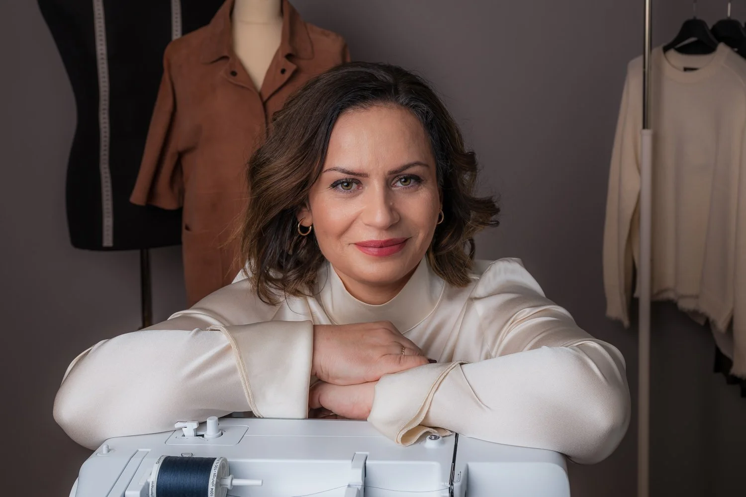 Eine Frau mit braunen lockigen Haaren, in einem cremefarbenen Satinoberteil, sitzt an einer Nähmaschine in einem Schneideratelier. Im Hintergrund sind Schneiderpuppen mit Kleidungsstücken und Bekleidung auf Kleiderstangen zu sehen.