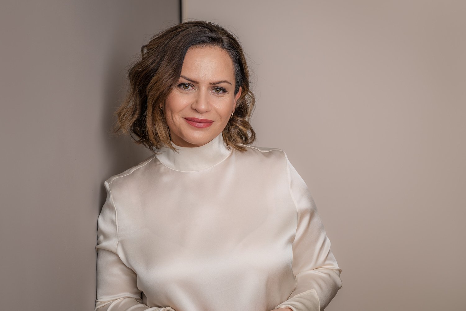Eine Frau mit schulterlangem, gewelltem braunem Haar, die einen weißen Satinblusen trägt, steht vor einer beige Wand und lächelt leicht.