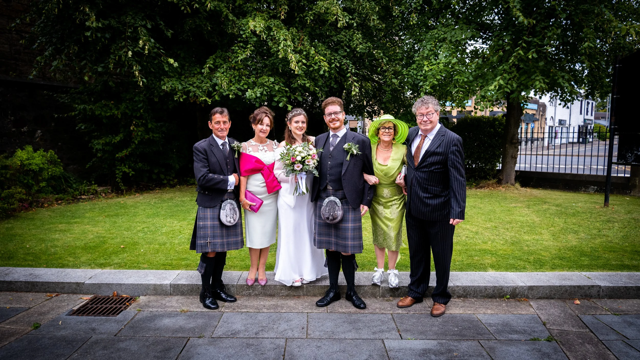 Group of six people at a wedding, standing on a sidewalk in front of a lawn and trees, dressed in formal and colorful attire.