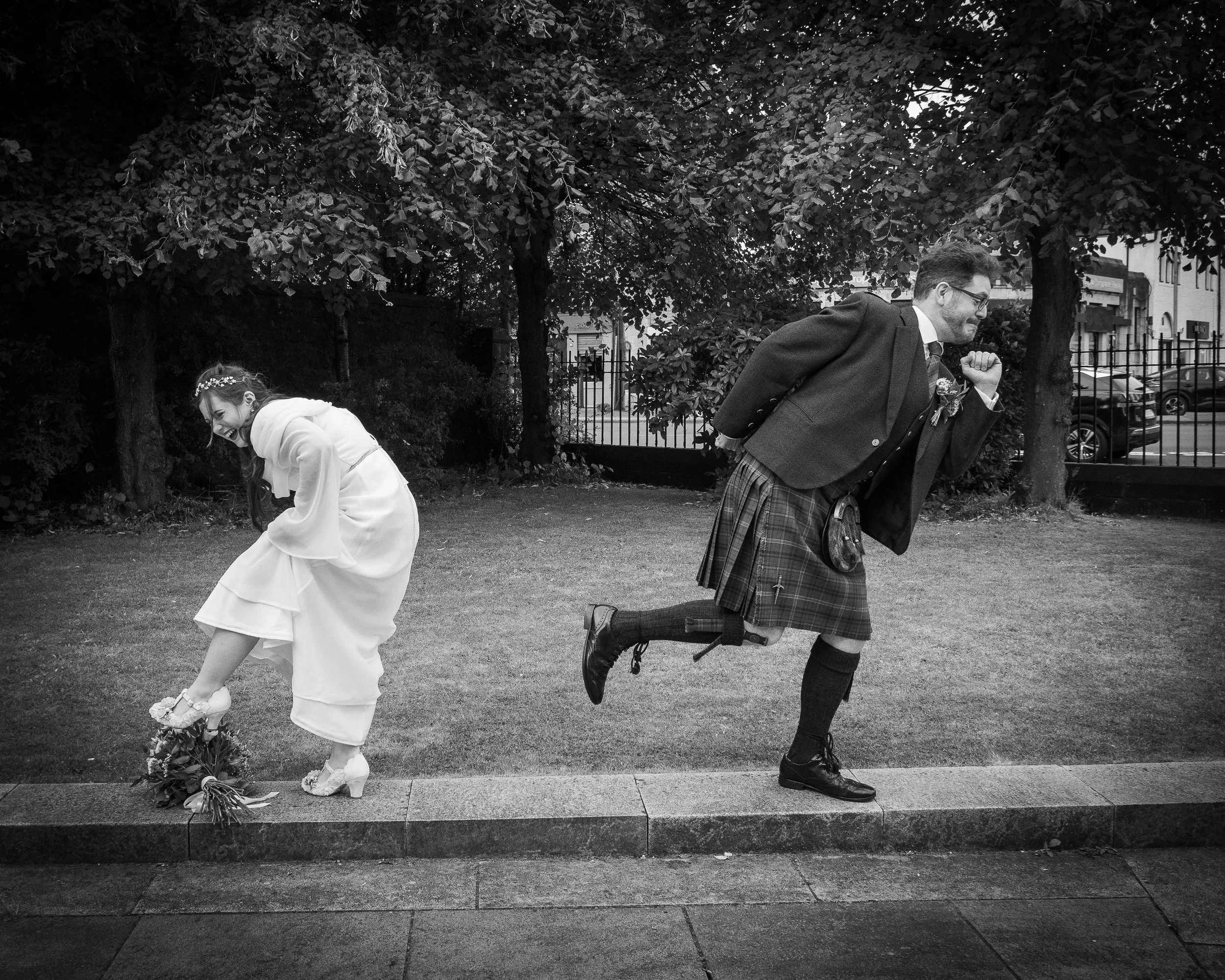 A black and white photo of a bride and groom outside. The bride is bending down, holding a bouquet, and laughing. The groom is playfully running behind her, dressed in traditional Scottish attire, including a kilt, jacket, and knee-high socks, with a