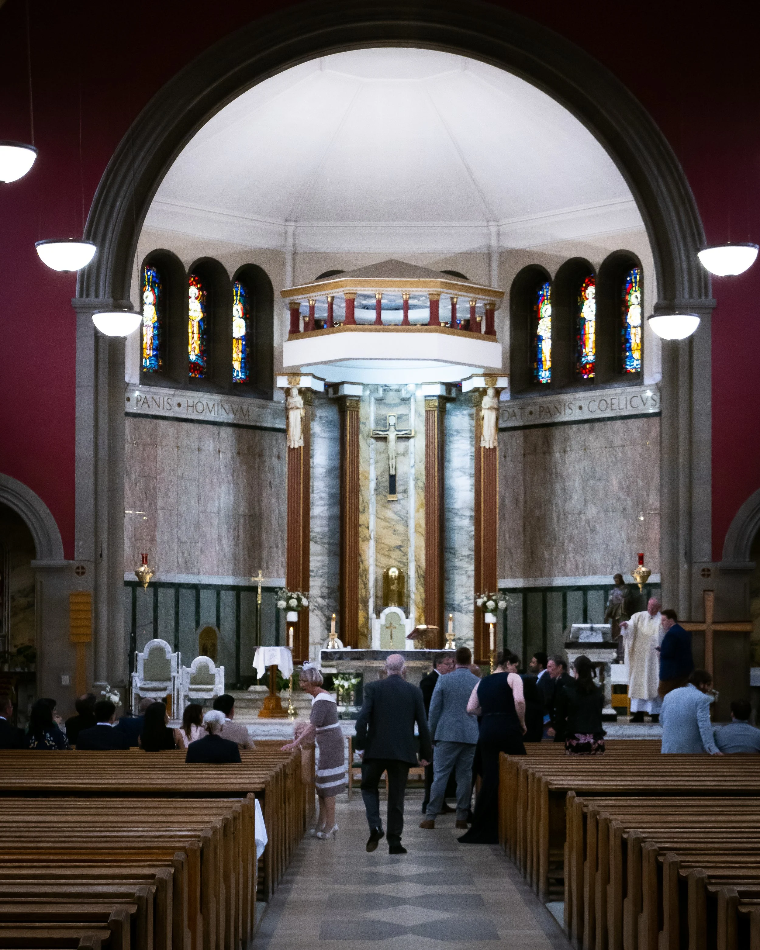 Interior of a church with people gathered at the altar area, wooden pews, stained glass windows, and a high arched ceiling.