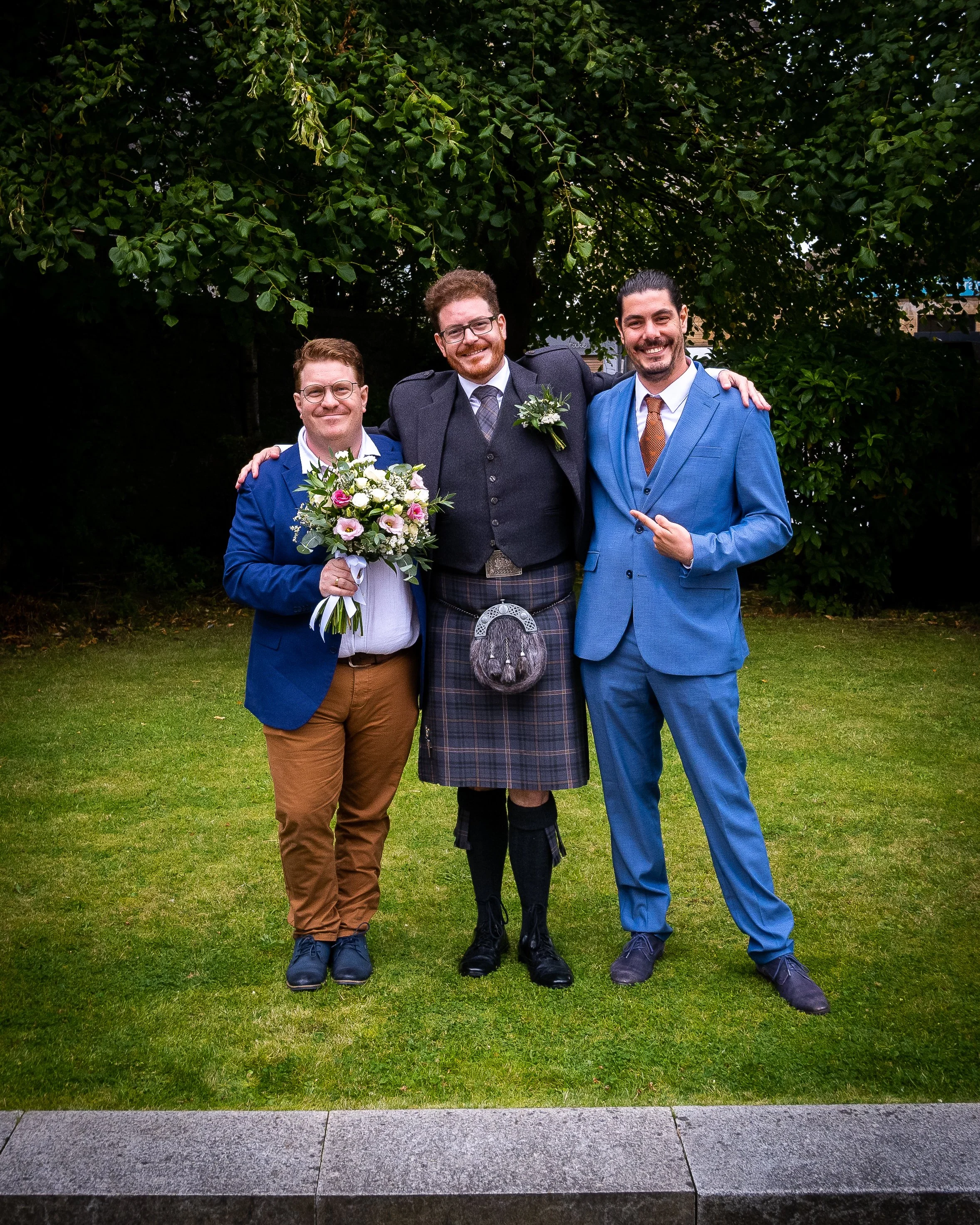 Three men standing outdoors on grass, dressed in formal attire, smiling, with lush green trees in the background. The man in the middle is wearing a kilt and holding a sporran, while the man on the left holds a bouquet of flowers.