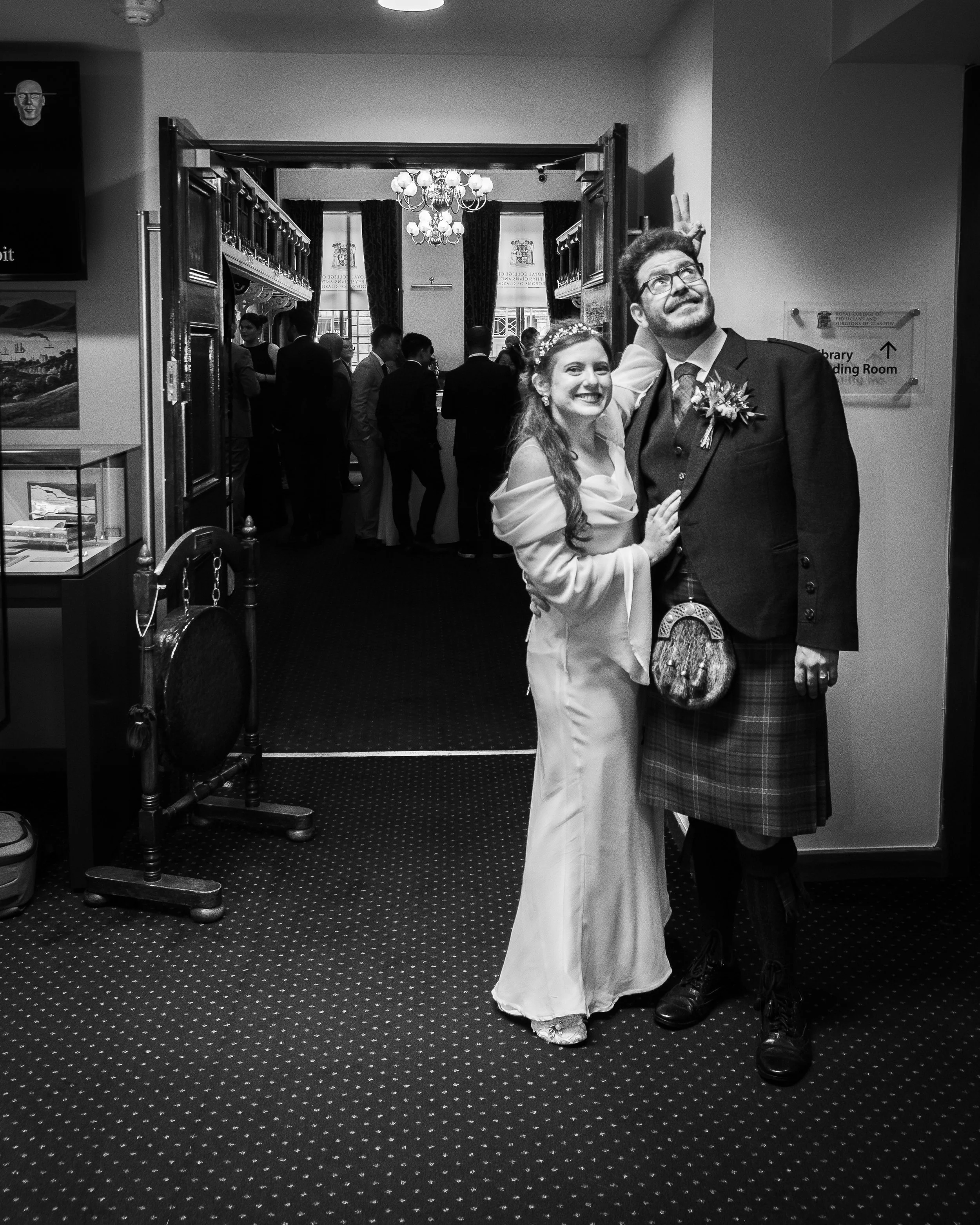 Bride and groom posing playfully at their wedding, with guests in the background inside a decorated venue.