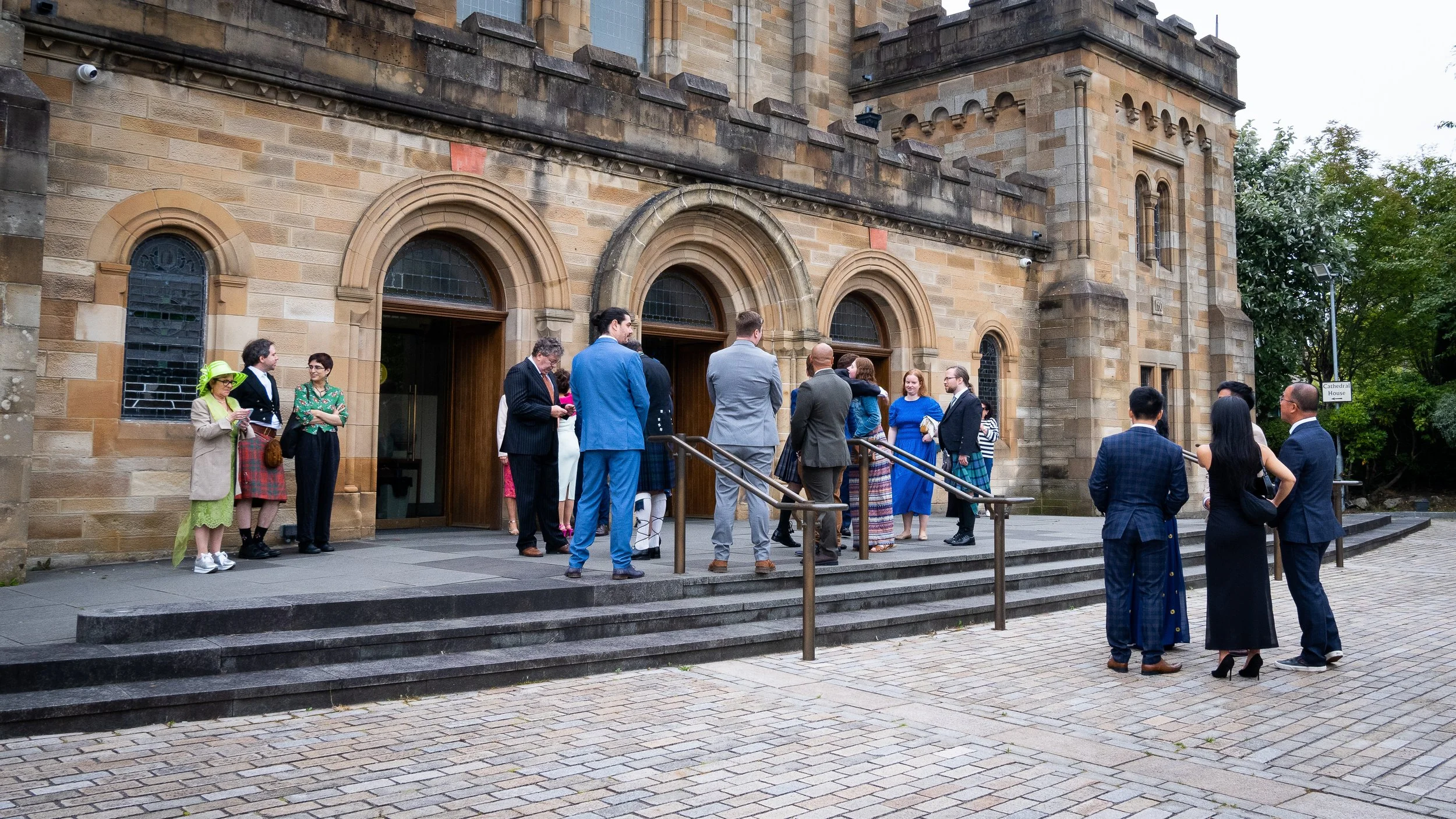 Group of people gathered outside a historic stone building, engaging in conversation, dressed in formal and semi-formal attire, with some women wearing hats, on a paved area with steps and a metal railing.