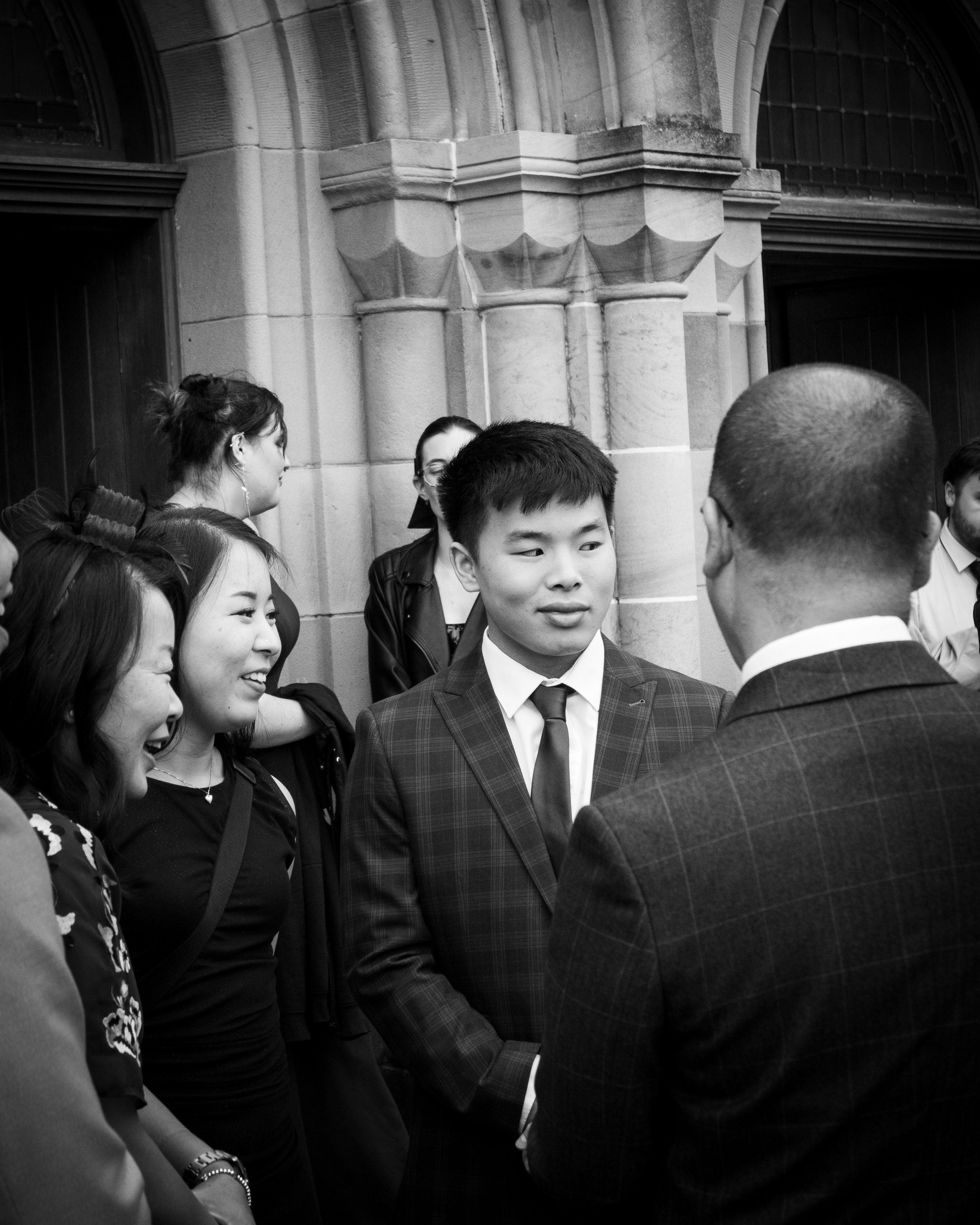 Group of diverse young adults in formal attire, engaged in conversation outside a stone building, some smiling and others listening.