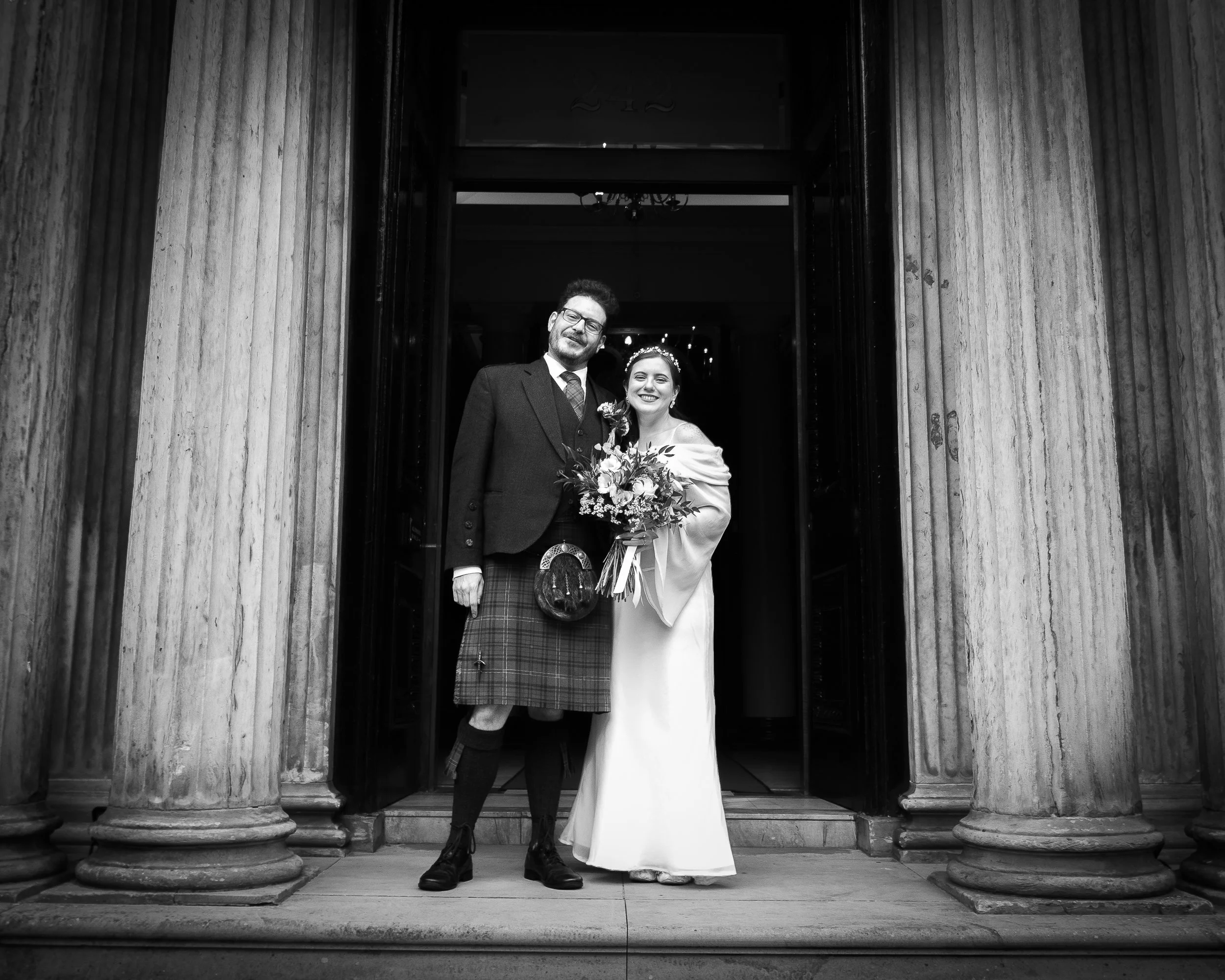 A black and white photo of a bride in a white gown and a groom in traditional Scottish attire standing at the entrance of a building with tall columns, smiling and holding a bouquet of flowers.