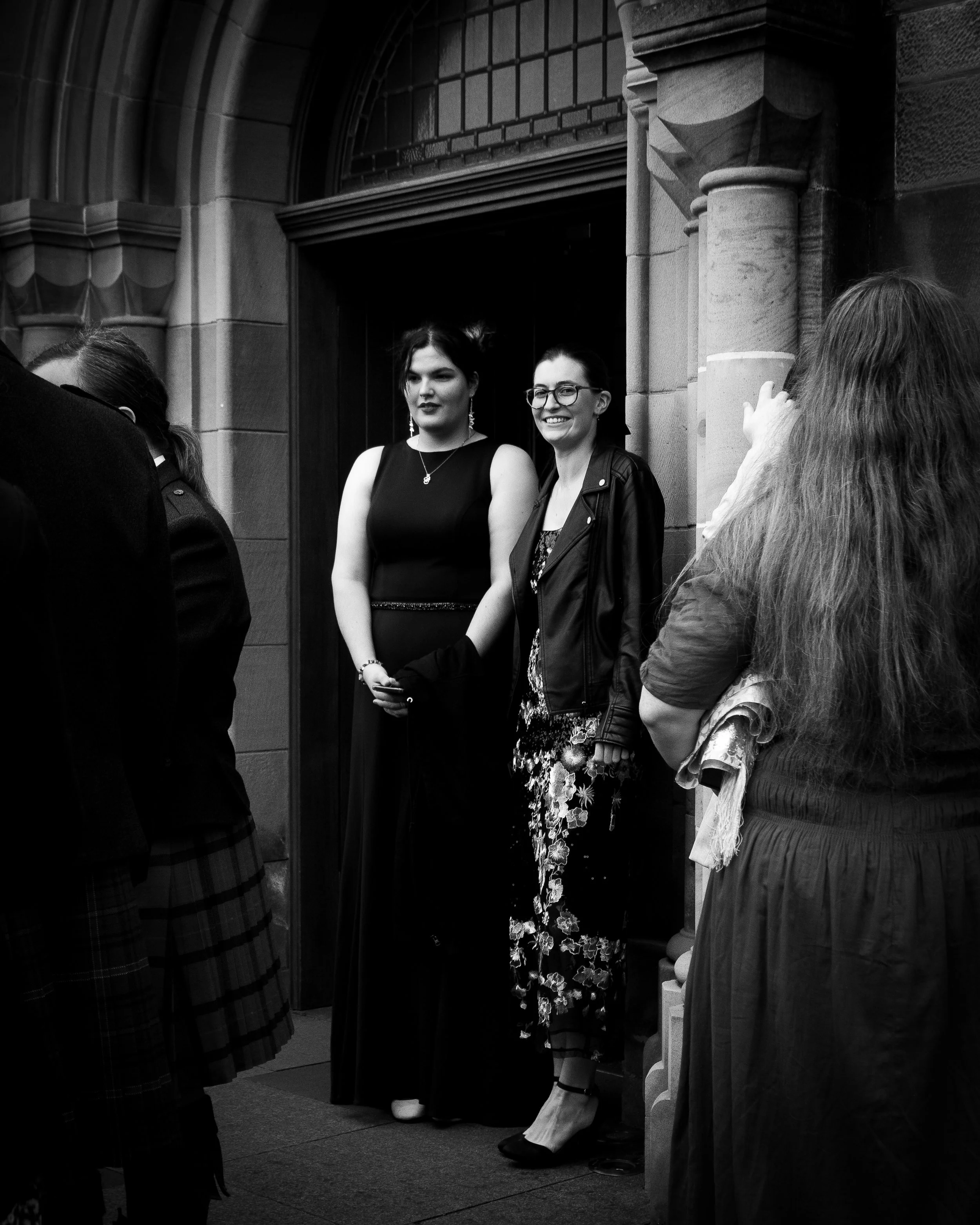 Two women standing together in front of a building entrance, smiling and posing for a photo, with other people around them.
