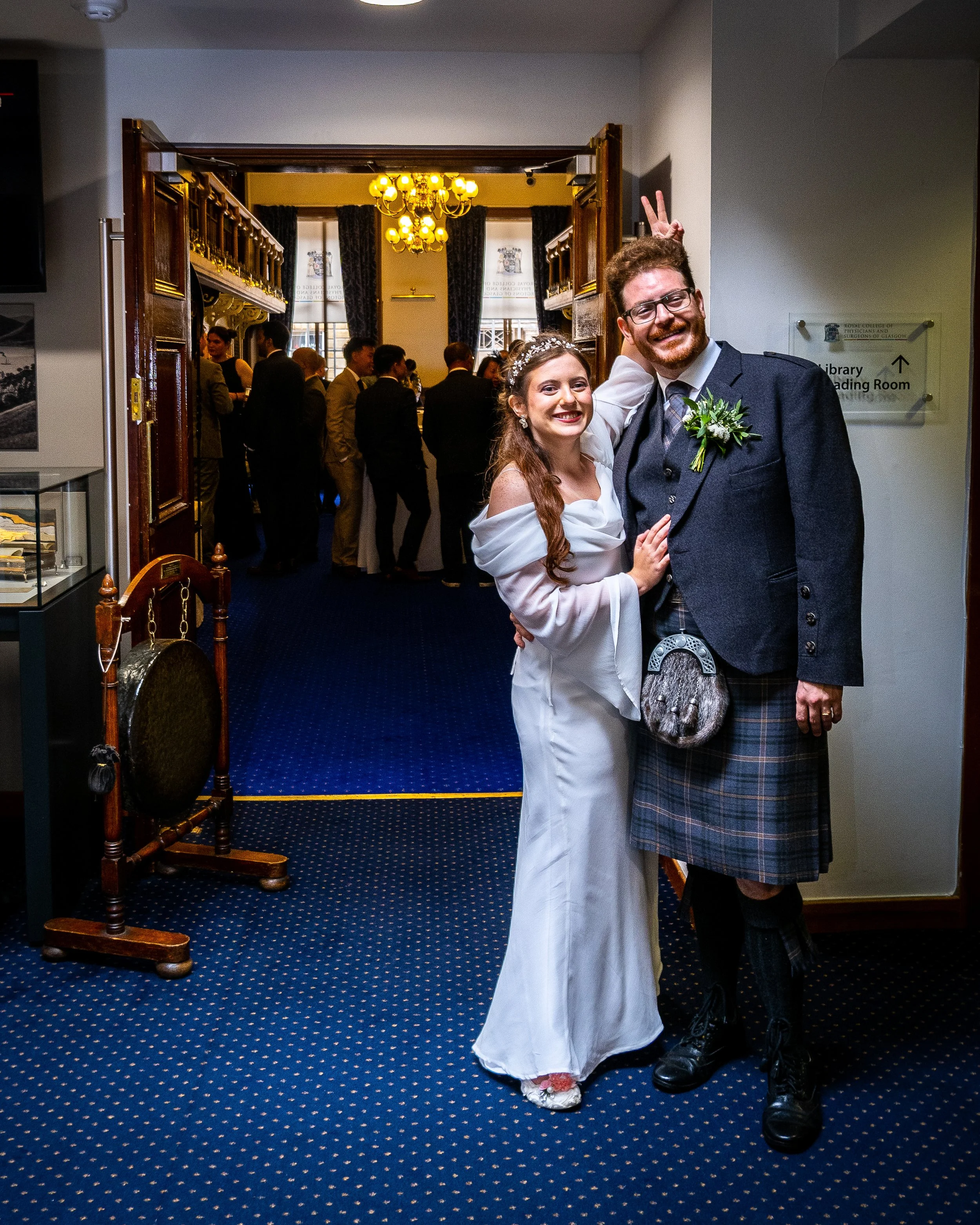 A smiling bride and groom in wedding attire posing together in a hallway with a blue carpet, with guests mingling in the background inside a decorated wedding venue.