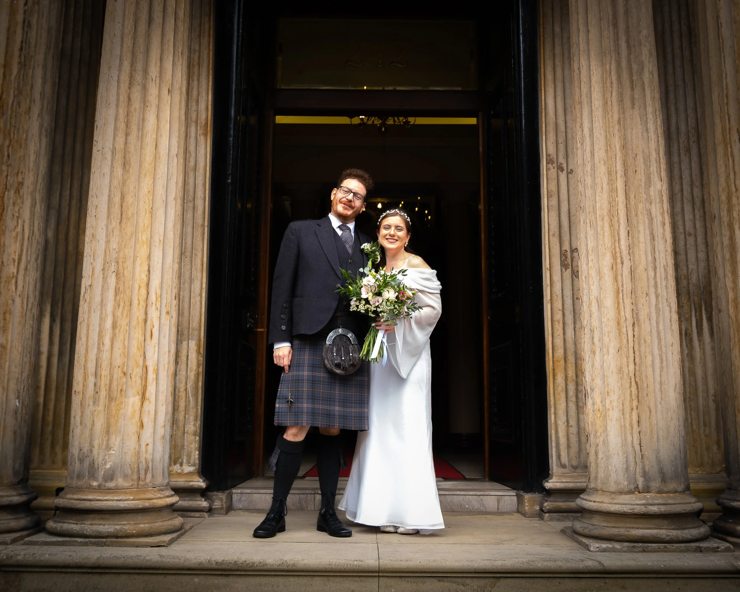 A bride and groom standing at the entrance of a building with large columns, smiling and holding a bouquet of flowers.