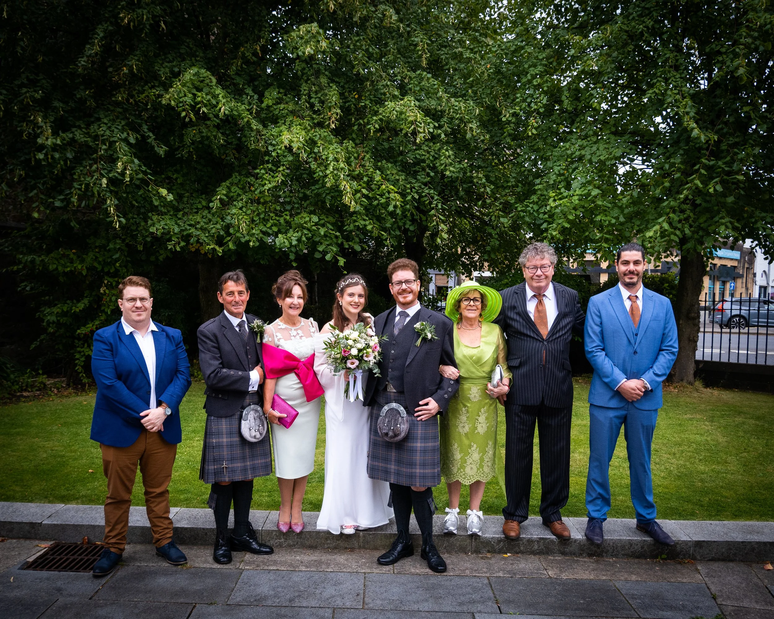 A group of nine people, including a bride and groom, standing outdoors on a lawn with trees in the background, dressed in formal wedding attire.