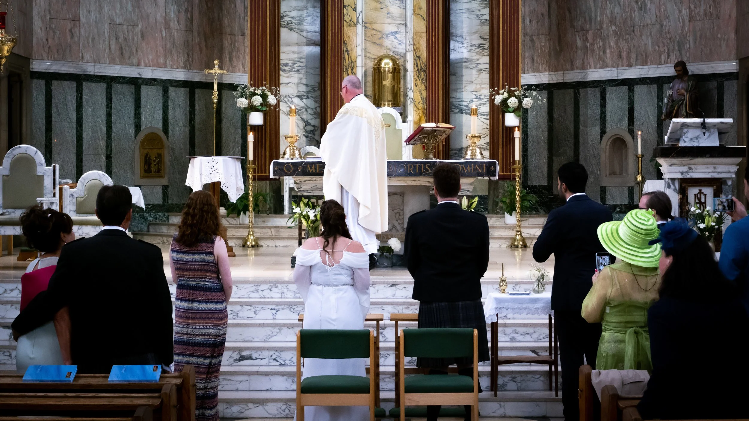 A priest leading a wedding ceremony in a church with seated guests, some taking photos, and decorative flowers and candles at the altar.