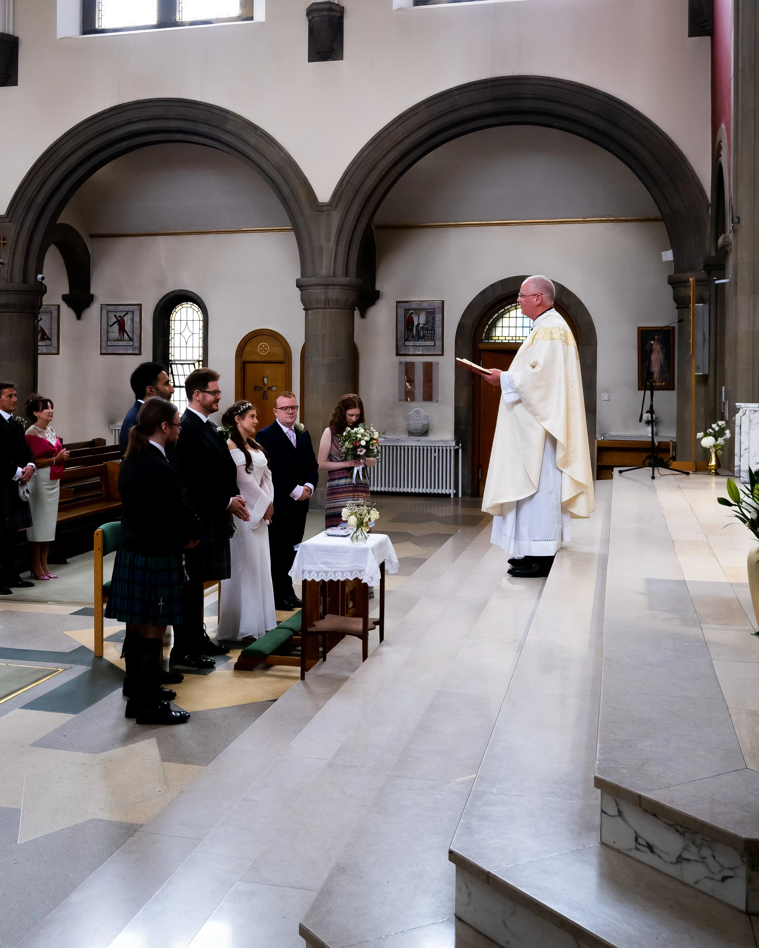 A wedding ceremony taking place in a church with clergy officiant and a group of formally dressed people, some in kilts, standing before the altar.