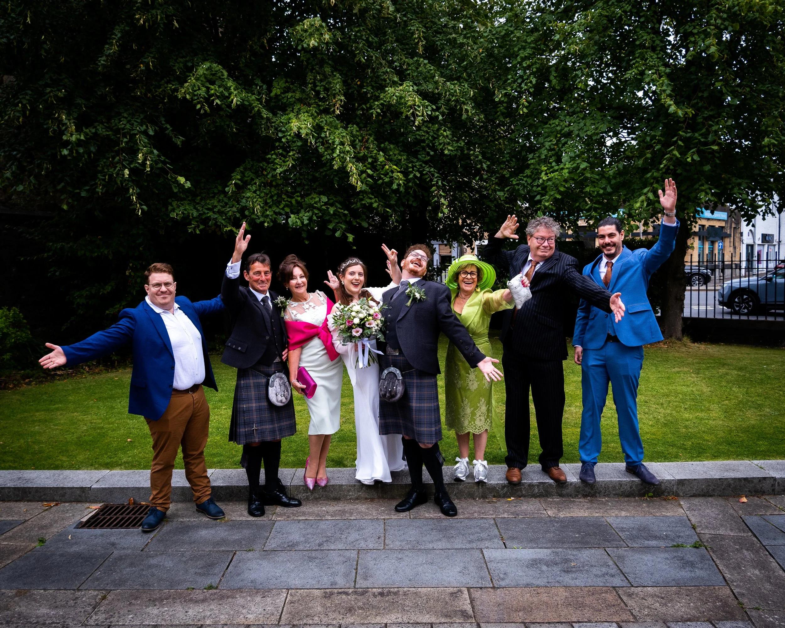 Group of nine people celebrating outdoors, including a bride with a bouquet, dressed in wedding attire, and others in colorful and formal clothing, standing on a sidewalk in front of a large green tree.
