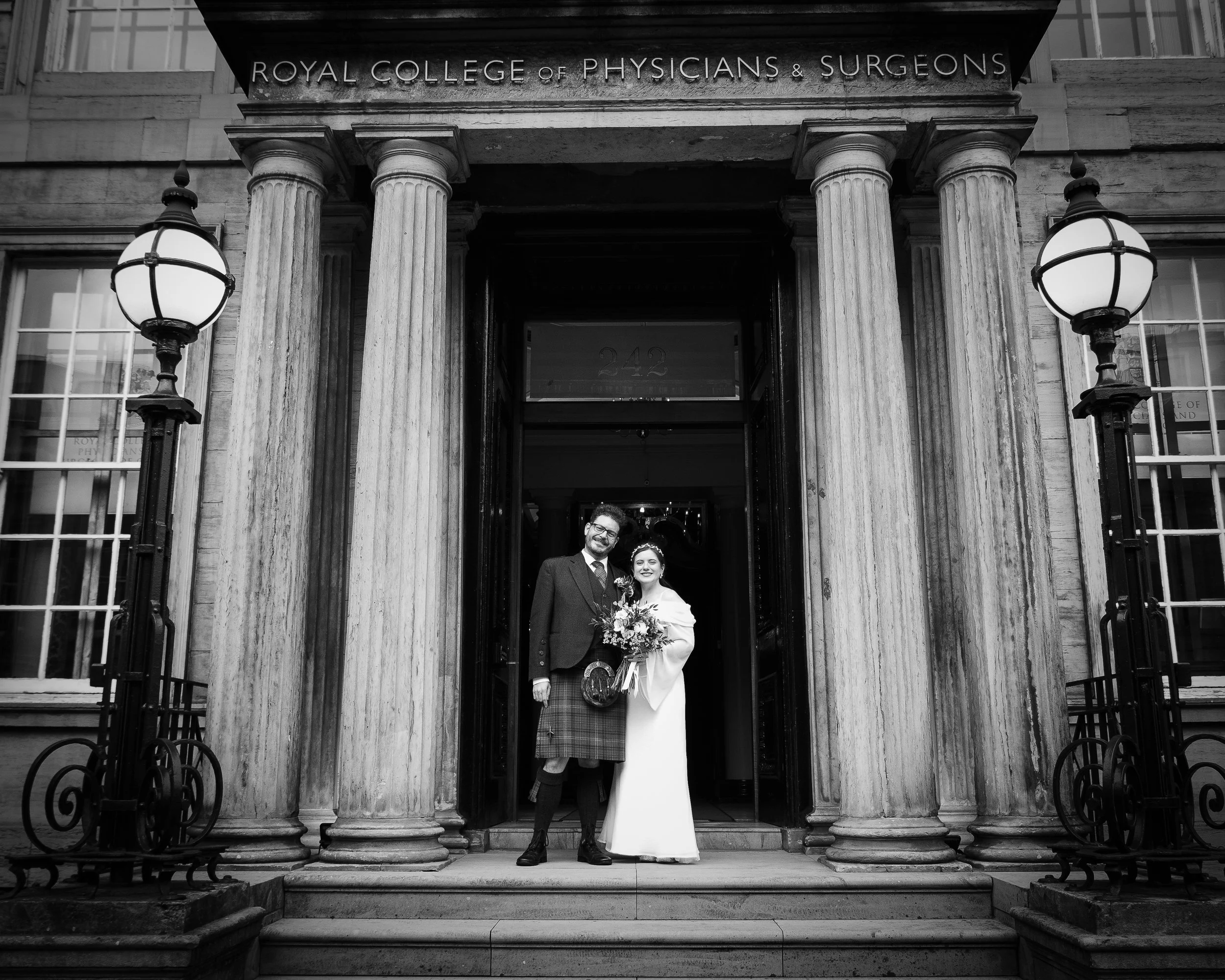 A black and white photograph of a newlywed couple standing beneath the entrance of the Royal College of Physicians & Surgeons, framed by four large columns with ornate capitals. The groom wears traditional Scottish attire, including a kilt and jacket