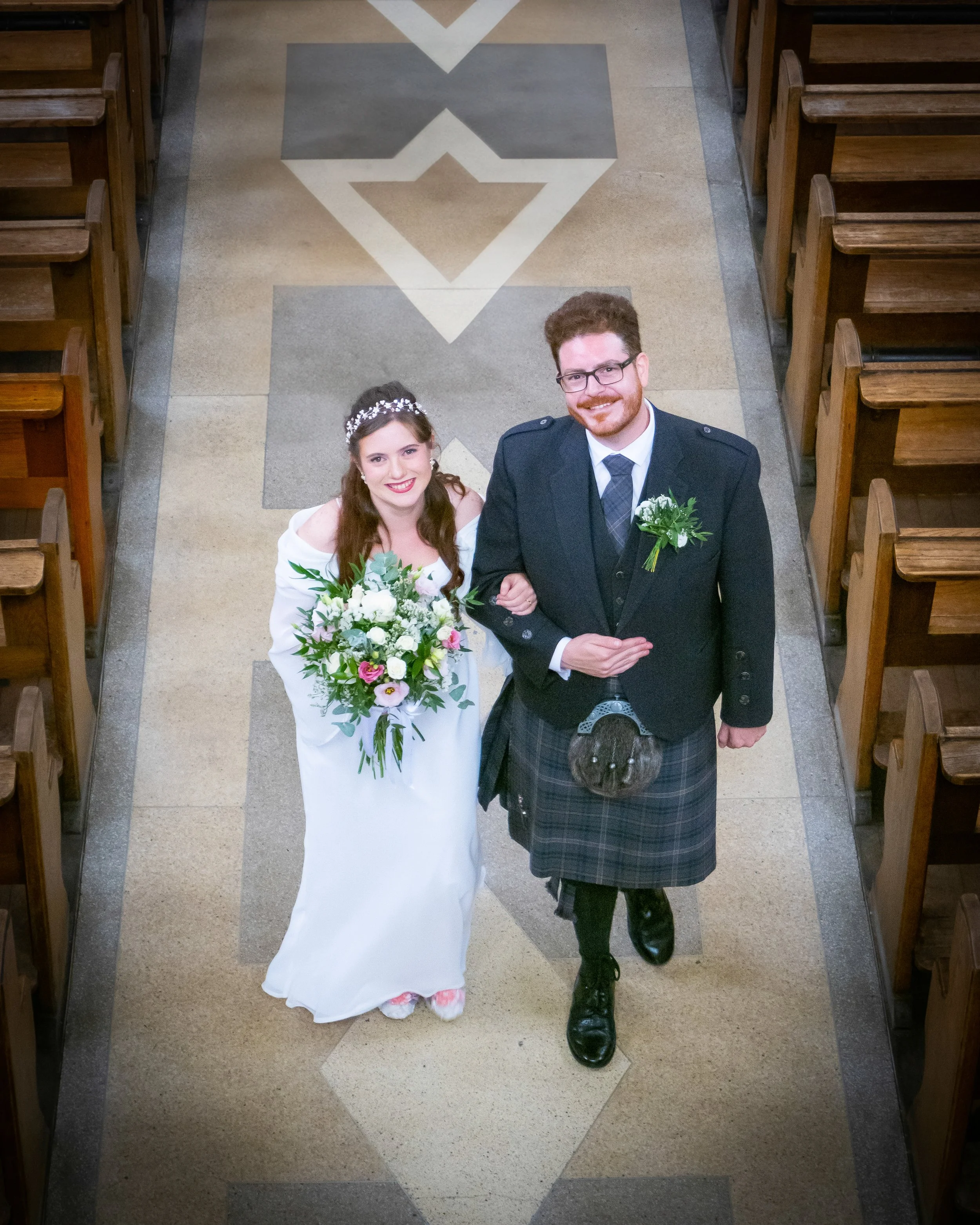 Overhead view of a bride and groom walking down the aisle in a church, with wooden pews on either side and decorative geometric patterns on the tile floor.