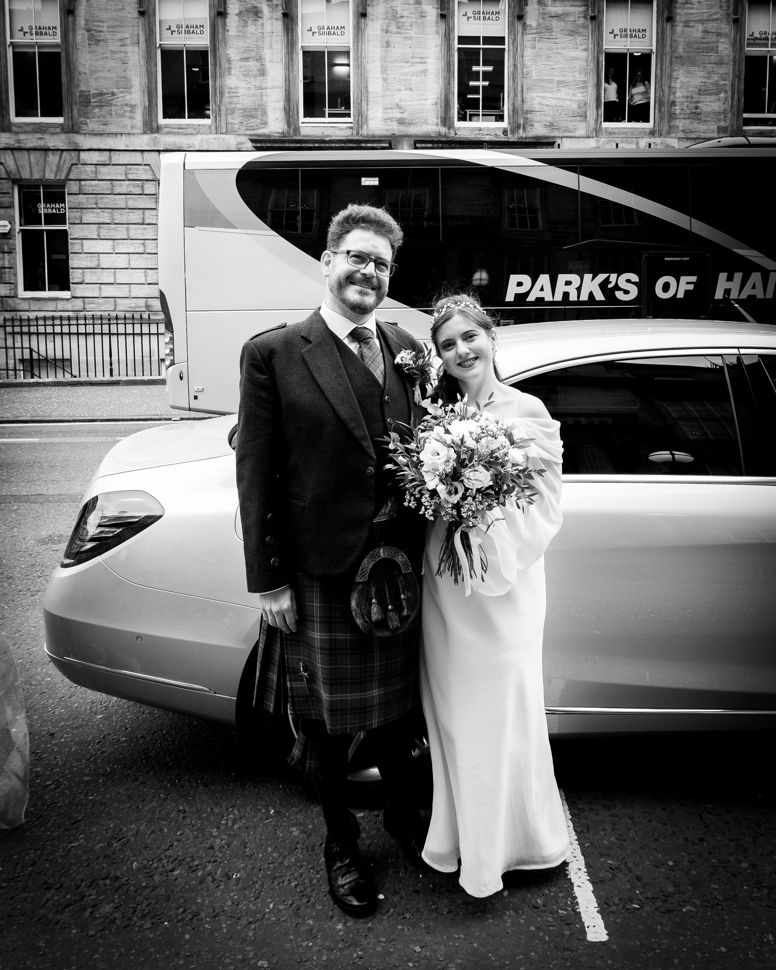 A groom in Scottish attire and a bride in a white wedding dress posing with a bouquet on a city street. Behind them are a car and a tour bus with 'Park's of H' visible on it. The scene is black and white.