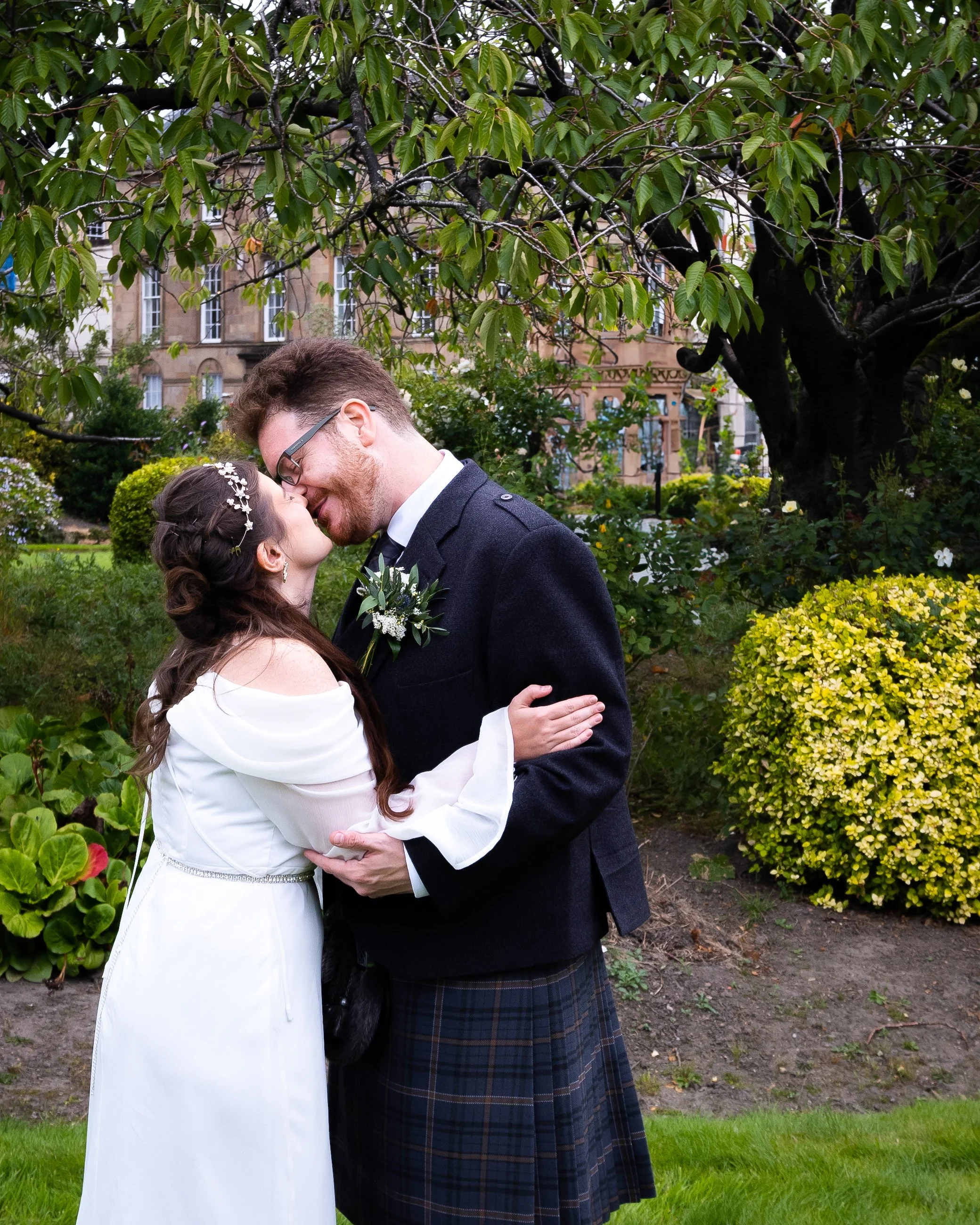 A bride and groom sharing a kiss outdoors in a garden, surrounded by greenery and trees, with a historic building in the background.