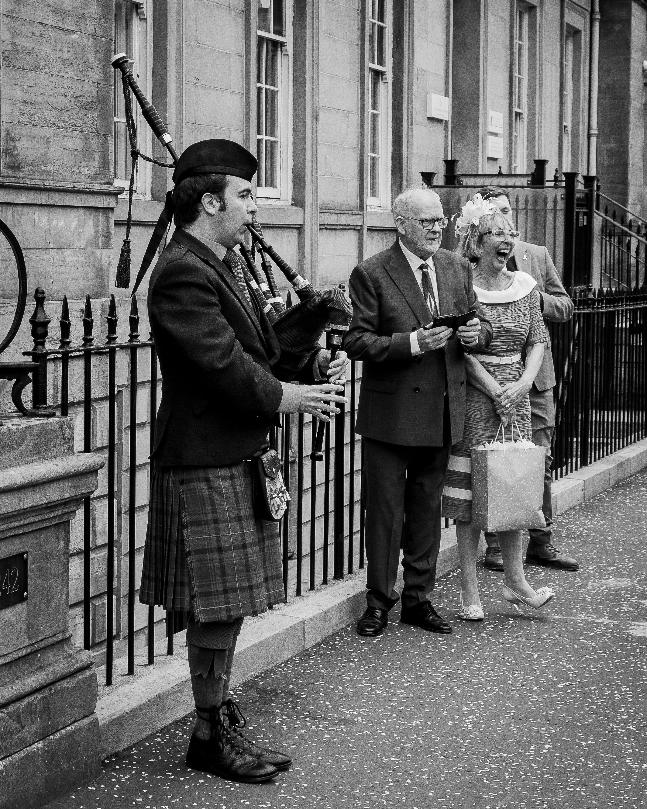 A man playing bagpipes standing on a city sidewalk, with three people laughing and watching nearby.