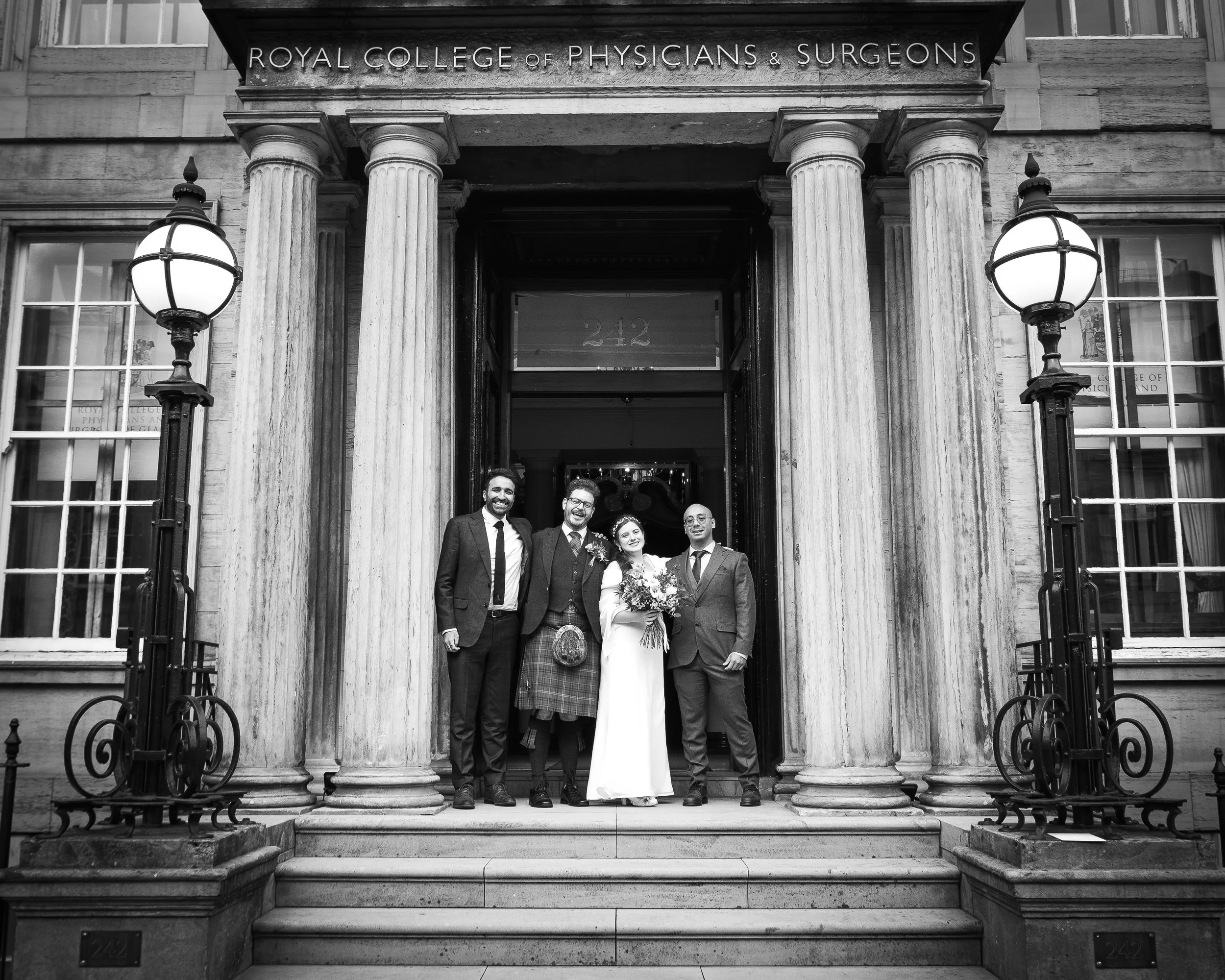 Four people dressed in formal attire standing on the steps outside the Royal College of Physicians and Surgeons building, with the bride holding a bouquet of flowers in the center.